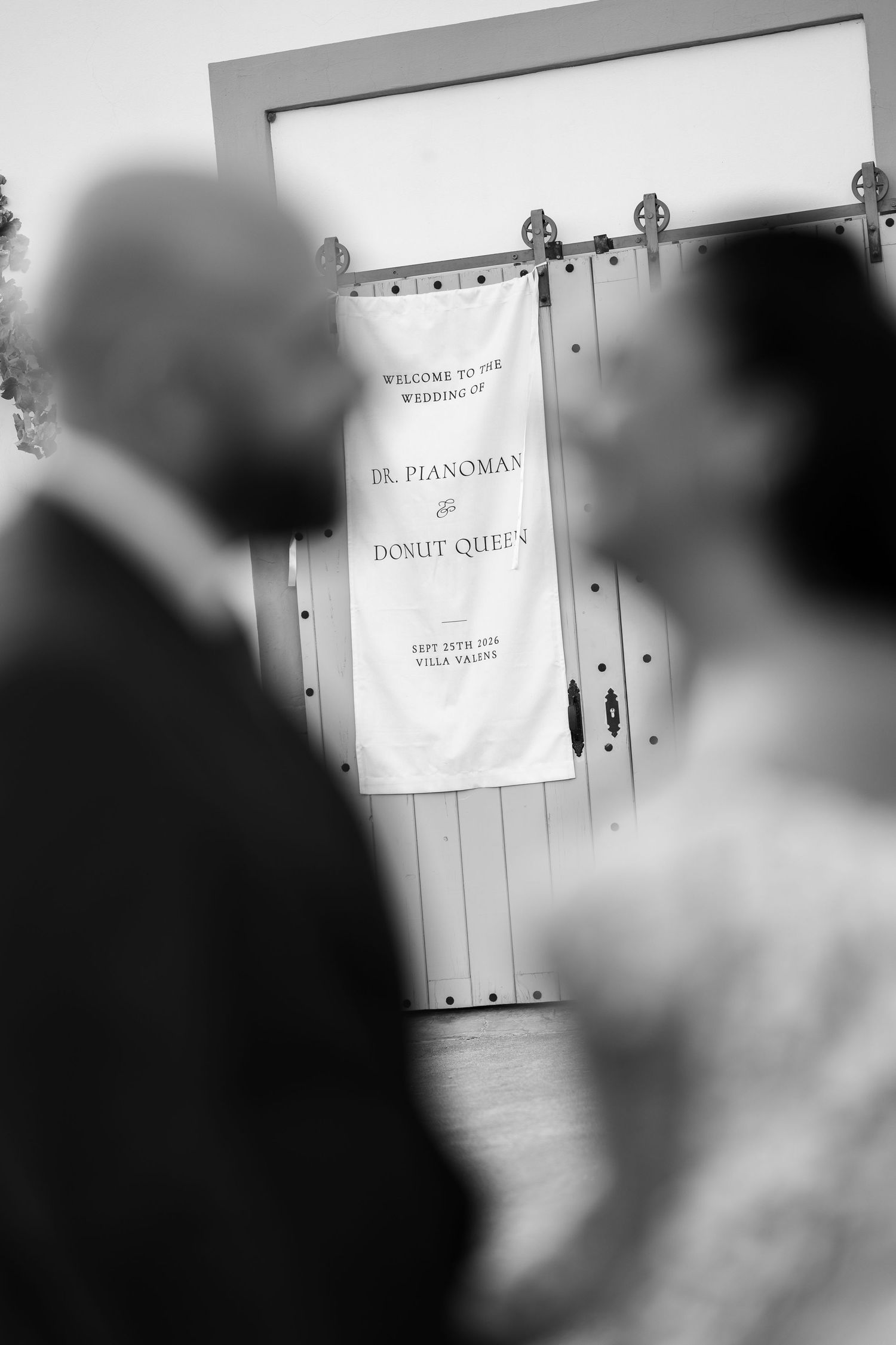Black and white photo of a wedding welcome banner reading Dr. Pianoman and Donut Queen, with two blurred guests in the foreground.