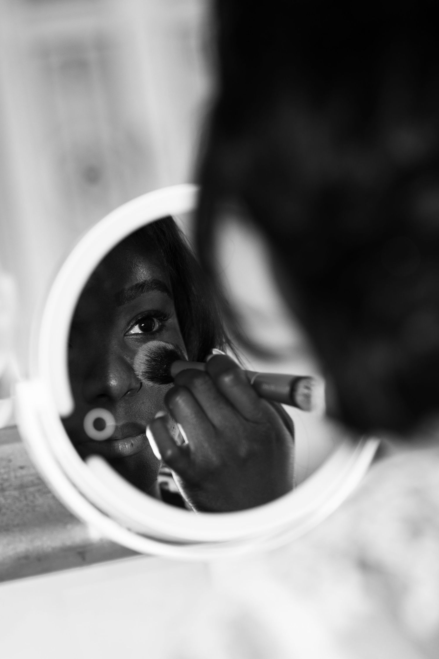Black and white photo of a woman applying makeup with a brush while looking into a round vanity mirror.