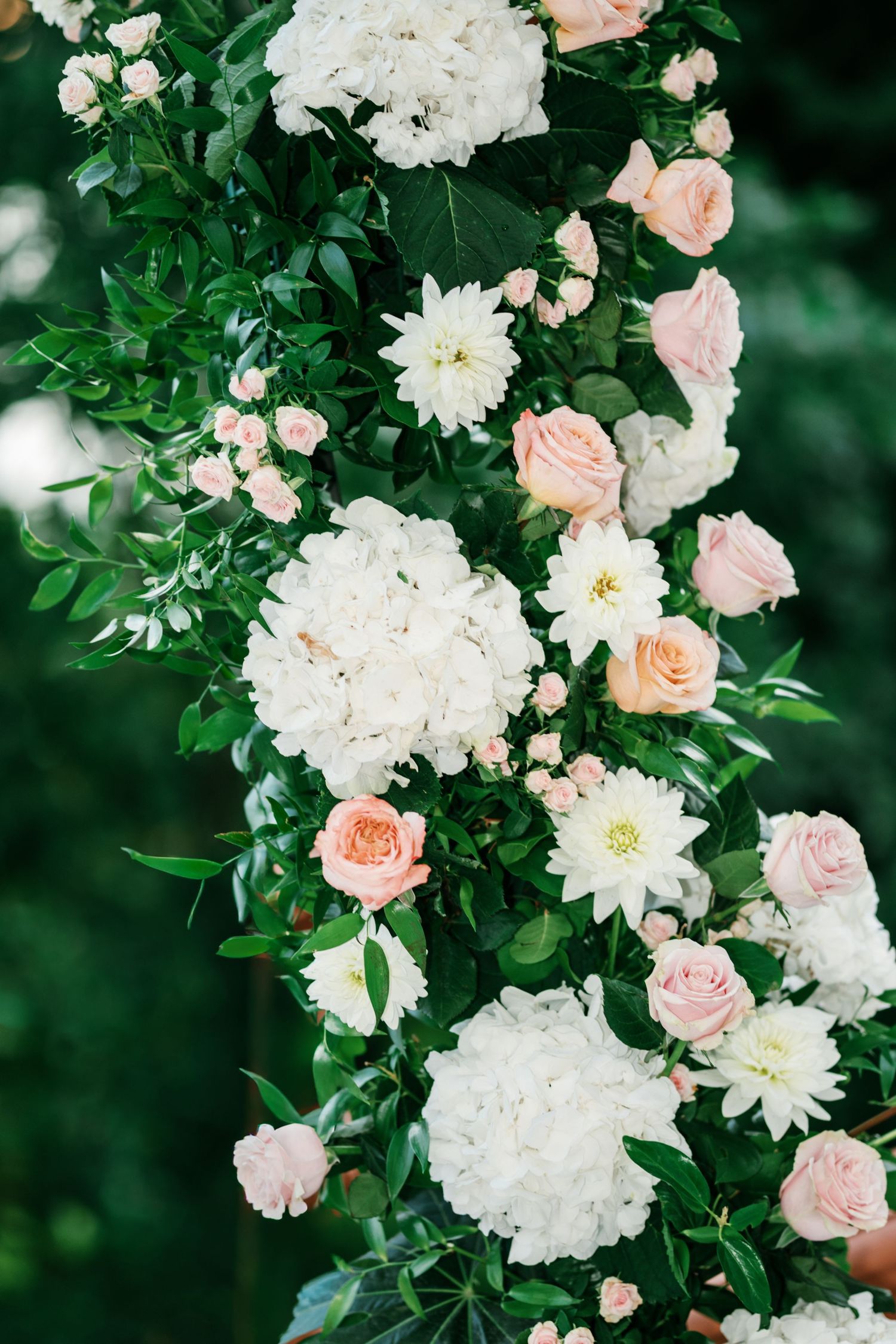 Wedding floral arrangement of white hydrangeas, blush pink roses, and lush green foliage against a soft blurred outdoor background.