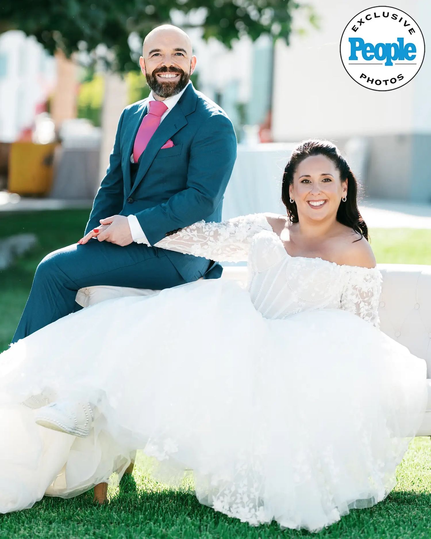 Smiling bride in an off-shoulder lace wedding gown sits beside a bearded groom in a teal suit and pink tie on a sunny lawn.