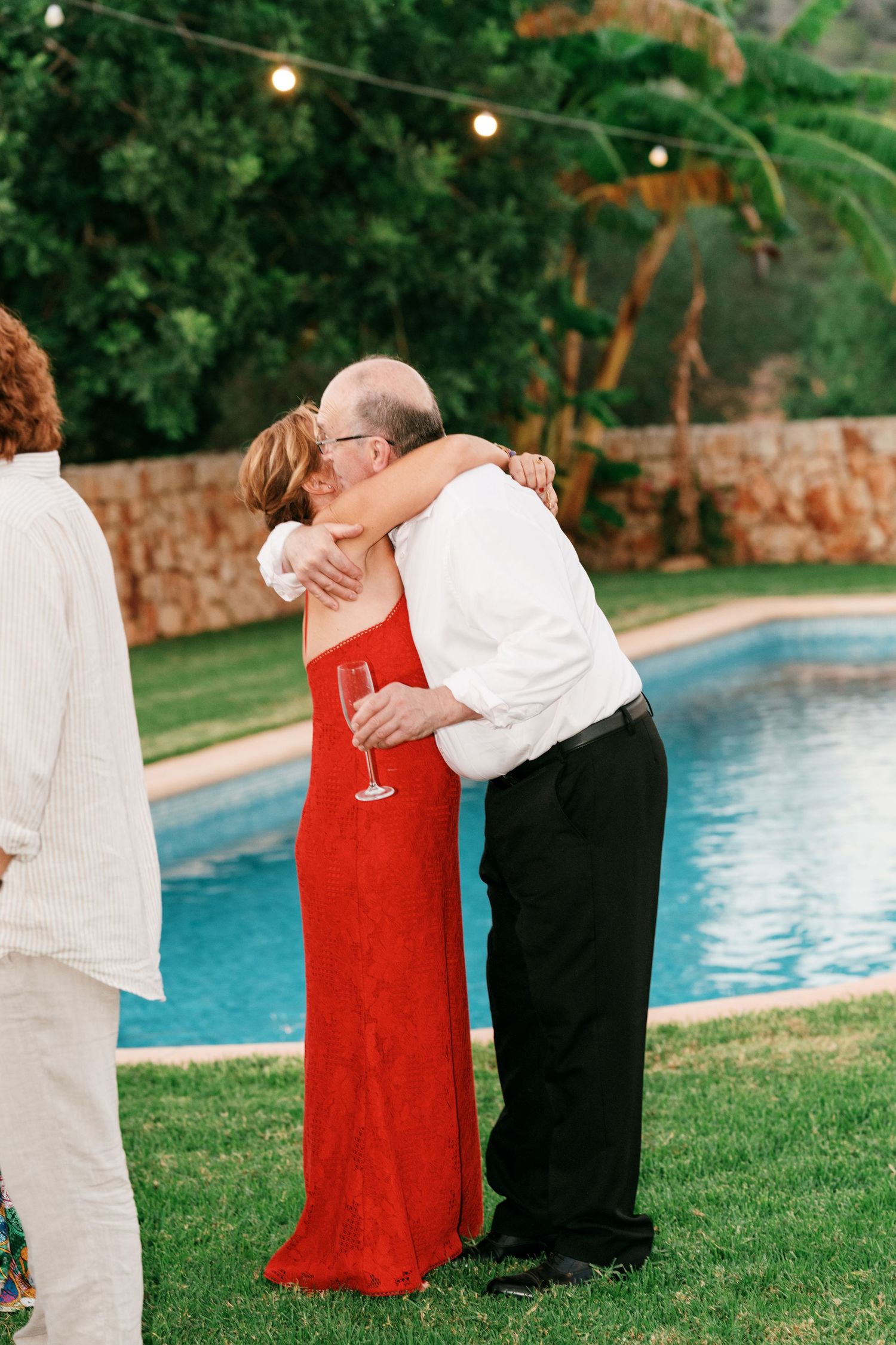 A woman in a red dress and a man in a white shirt embrace on a lawn beside a swimming pool under string lights at an evening party.