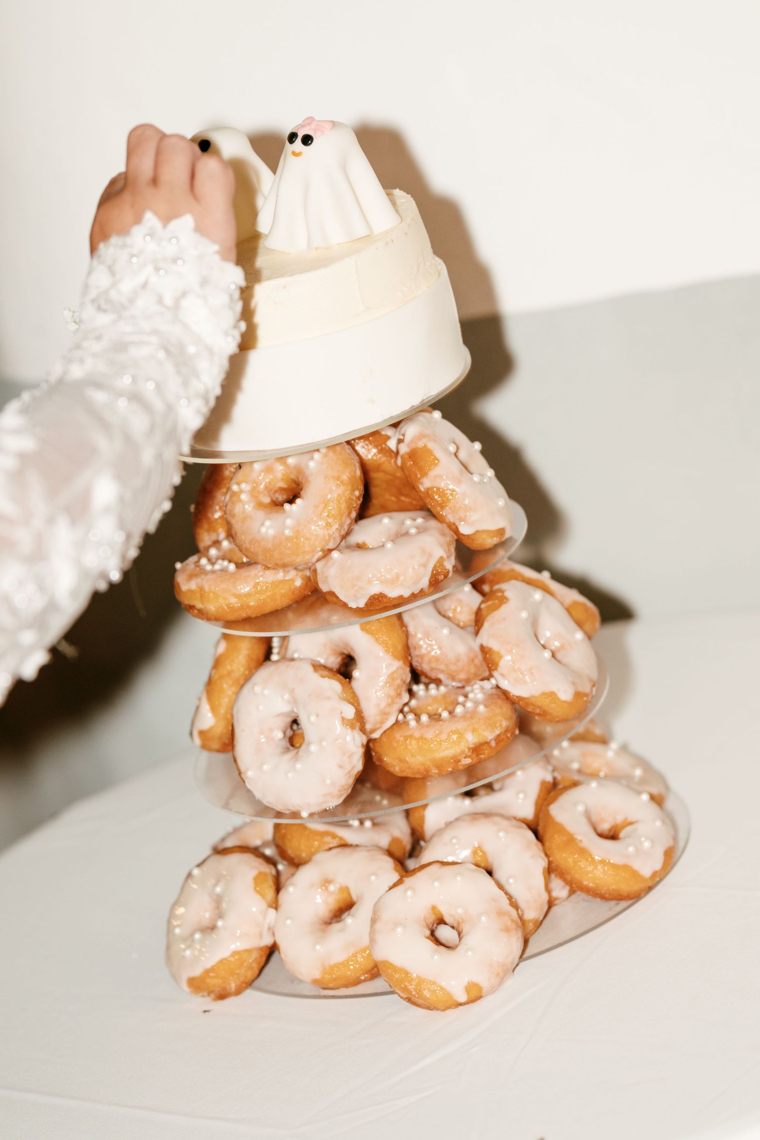 A tiered tower of glazed donuts with sprinkles topped by a small ghost cake topper, with a hand in a lace sleeve reaching toward it.
