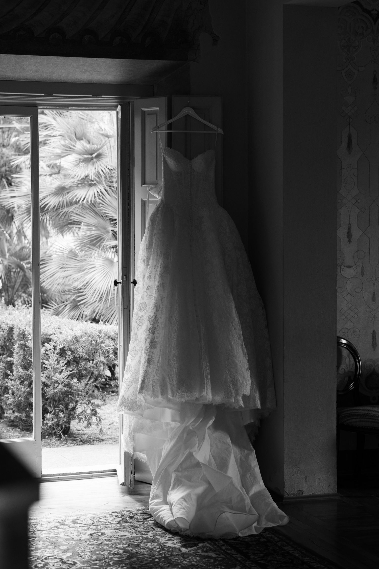 Black and white photo of a strapless lace wedding dress hanging from an open door with palm trees visible outside.