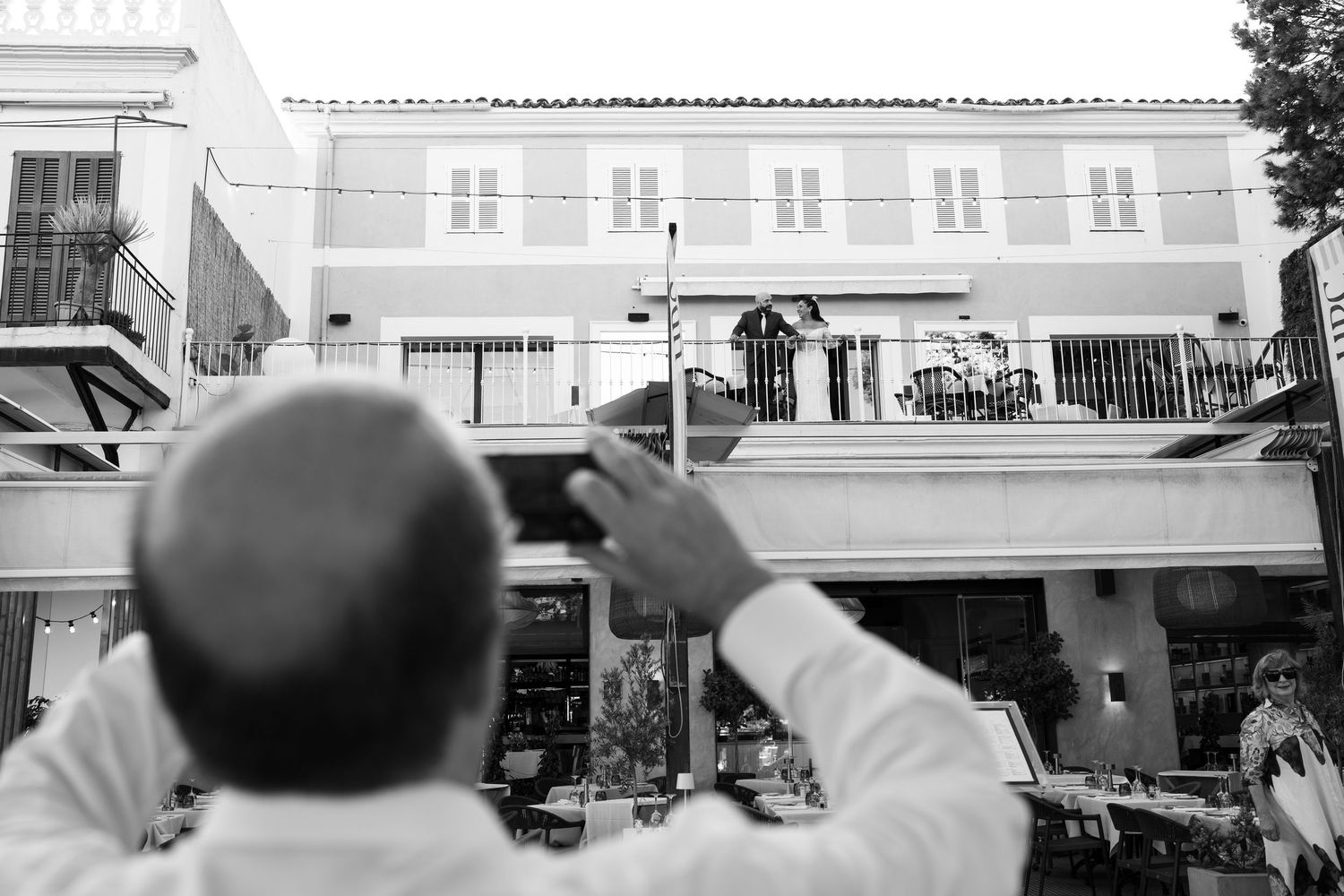 Black and white photo of a man from behind photographing a Mediterranean-style building with balconies and a figure on a terrace.