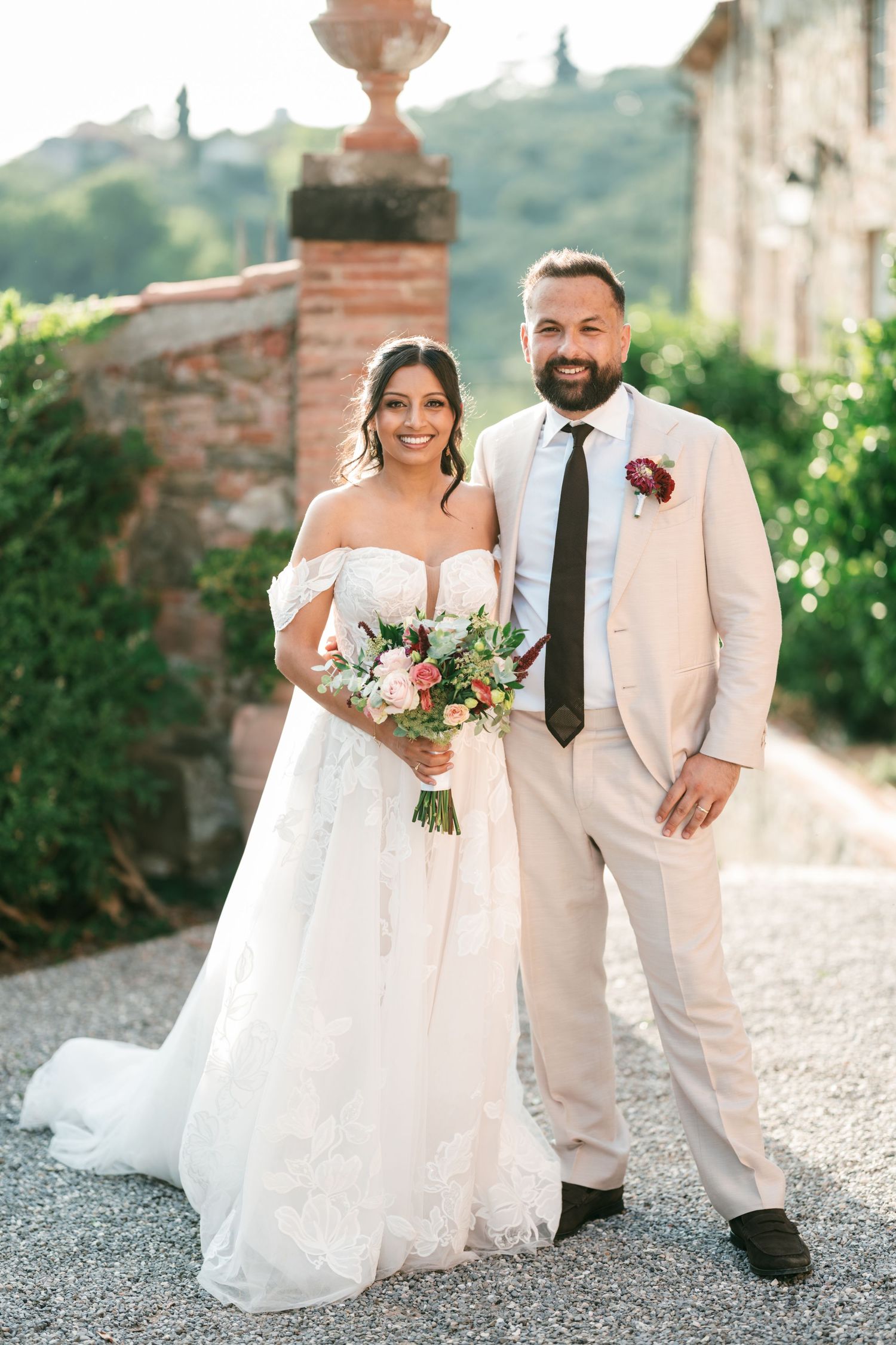 Smiling bride in an off-shoulder lace wedding gown holding a bouquet stands beside a groom in a beige suit on a gravel path.