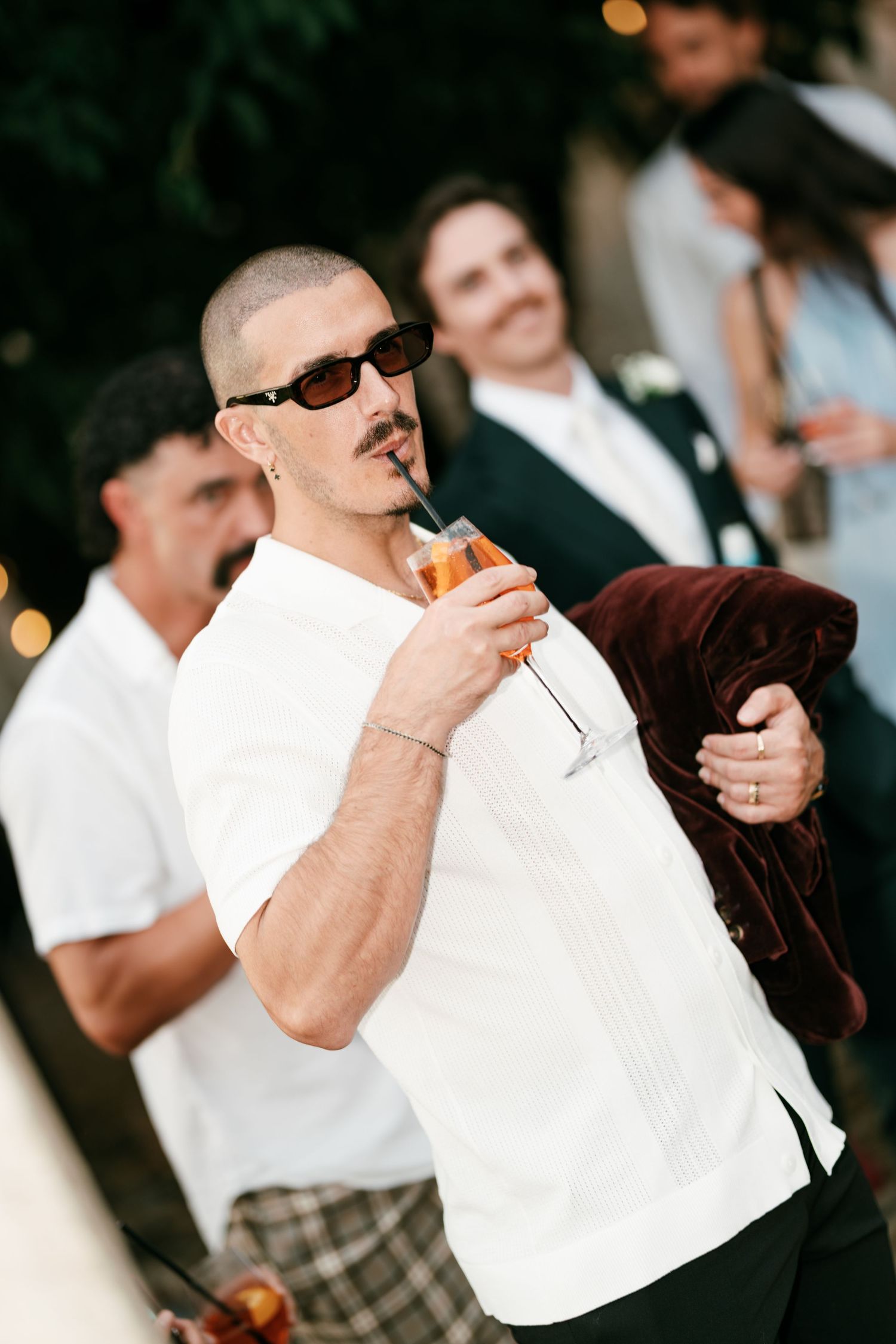 A man in sunglasses and a white guayabera shirt sips an orange cocktail at an outdoor wedding while guests chat behind him.