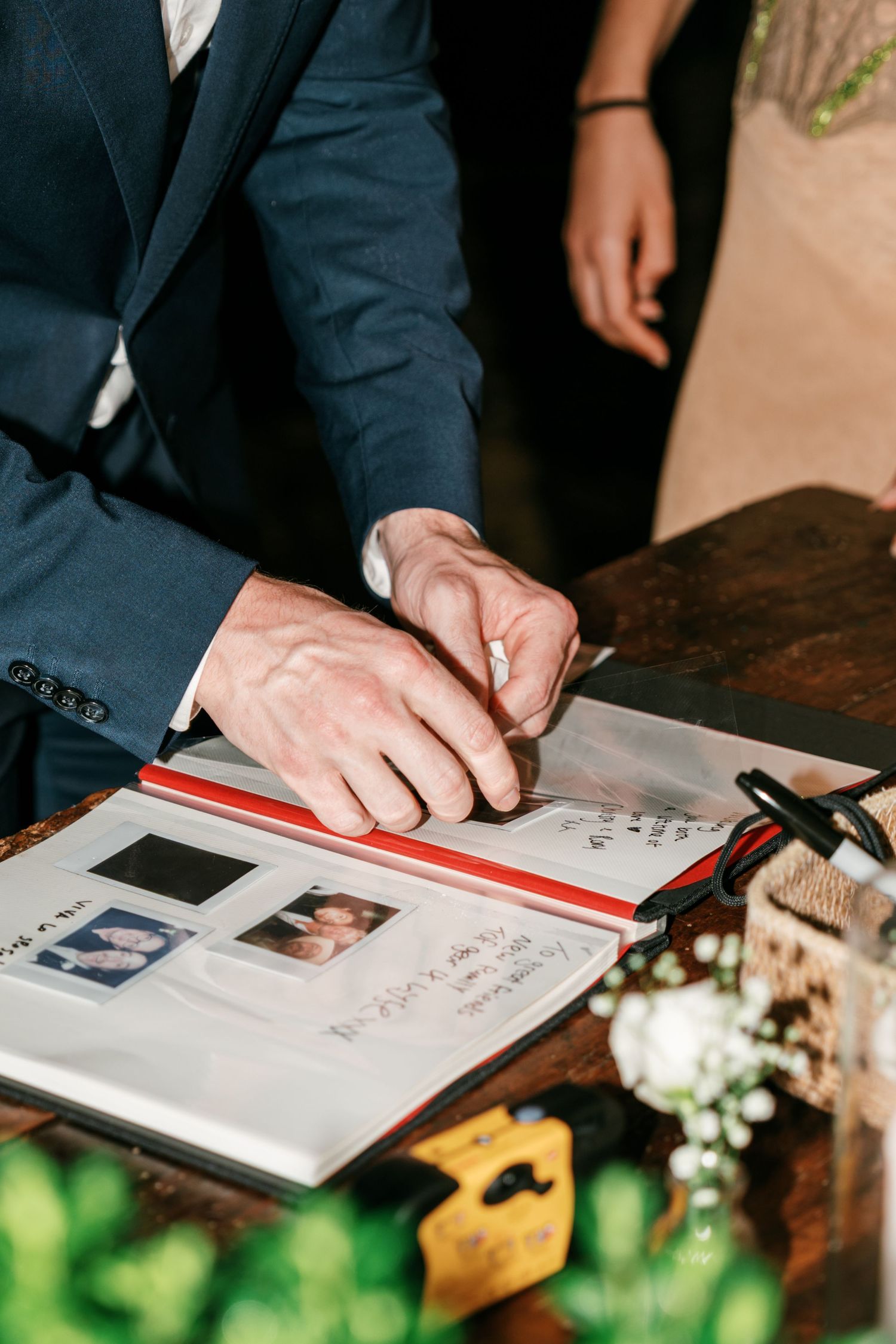 A groom in a navy suit signs a wedding guest book with photos and handwritten notes, beside a vase of white flowers.
