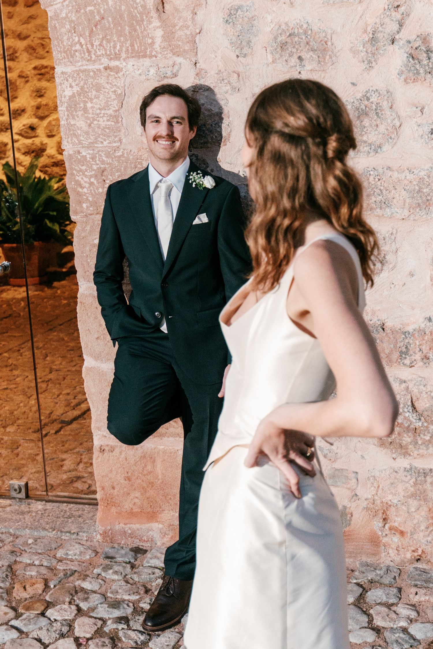 Smiling groom in a dark green suit with a boutonniere leans against a stone wall as the bride in a white satin dress looks on.