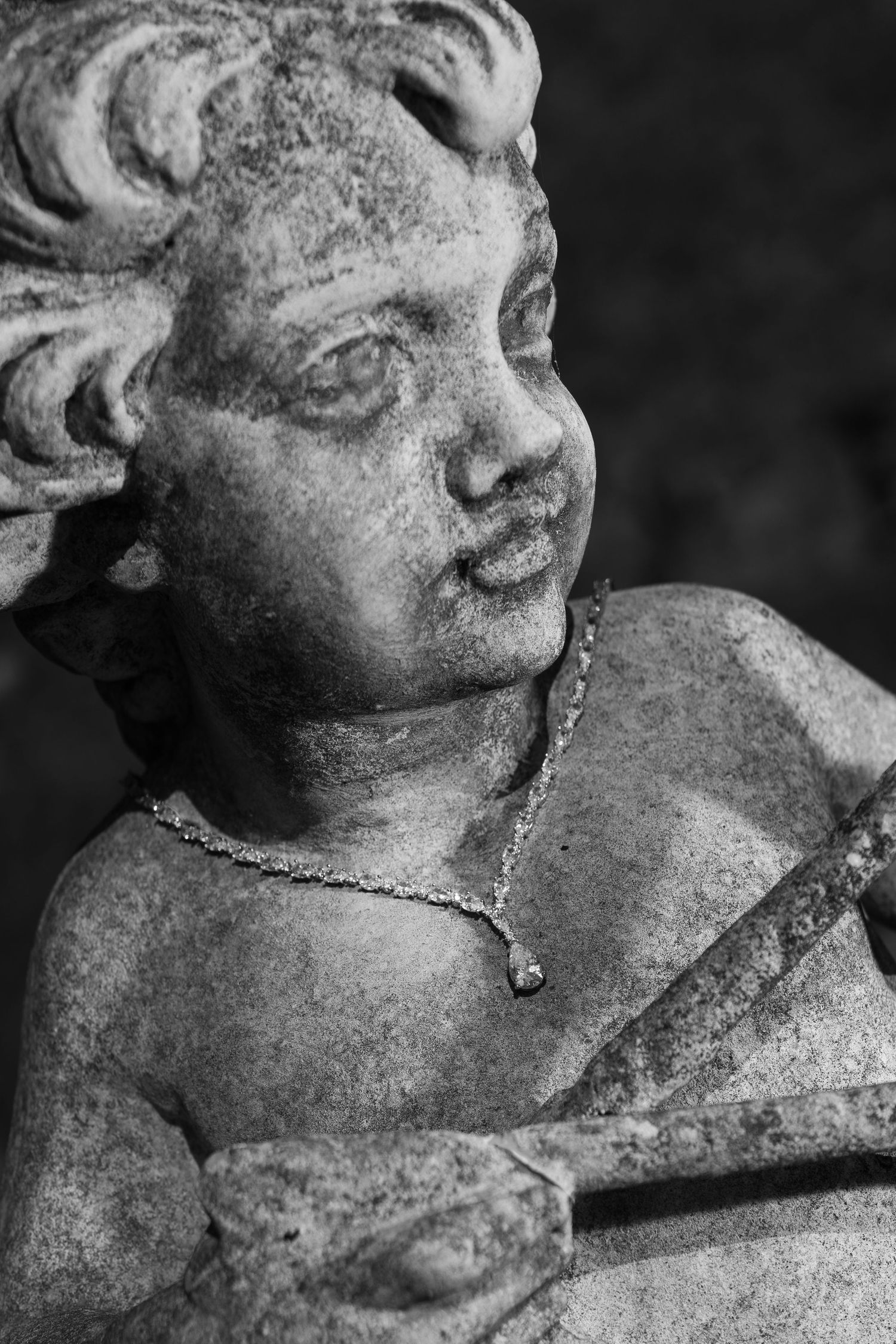 Black and white close-up of a weathered stone cherub statue with curly hair, a delicate pendant necklace, and a bow in its hands.