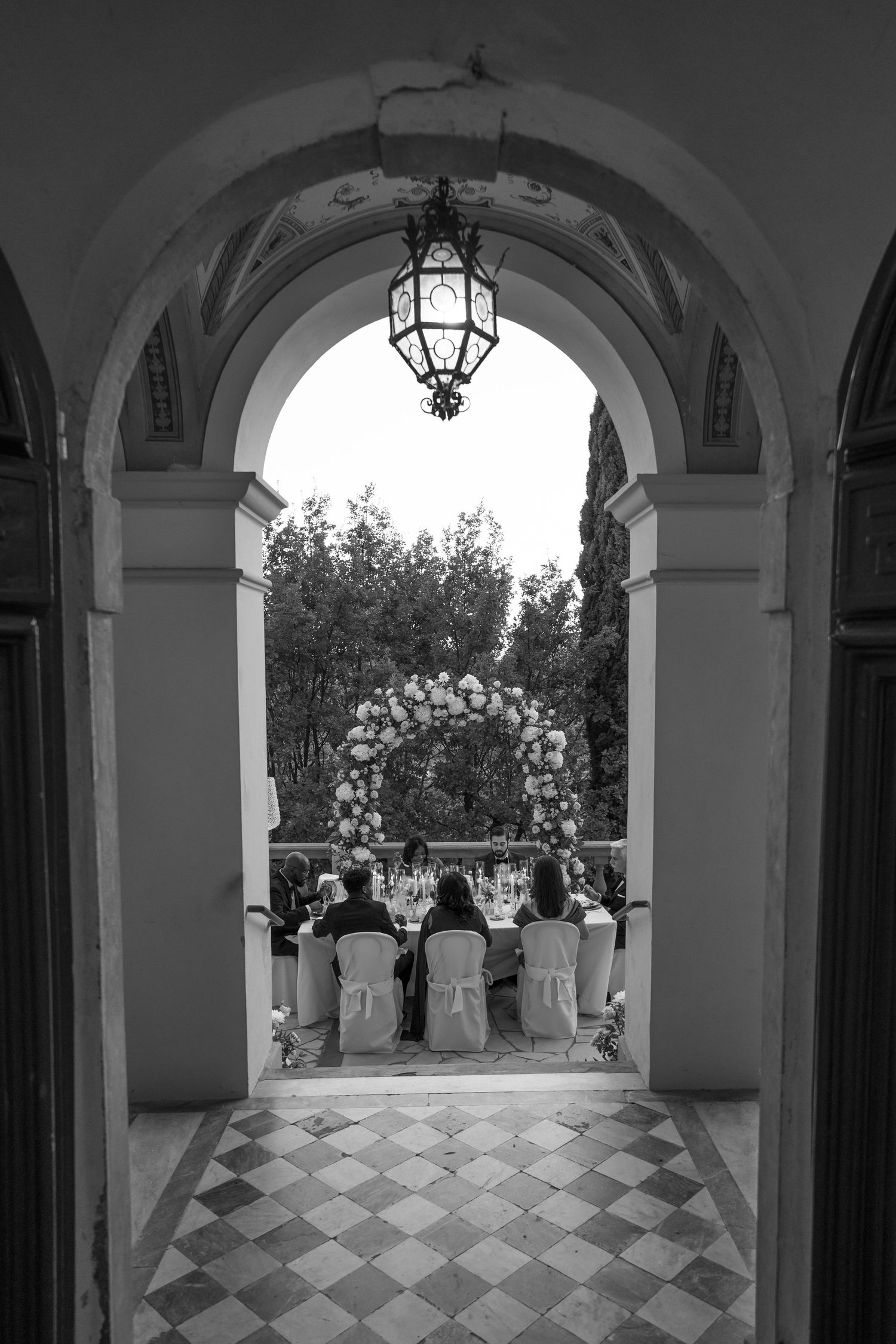 Black and white view through a stone archway of wedding guests seated at a reception table in front of a round floral arch.