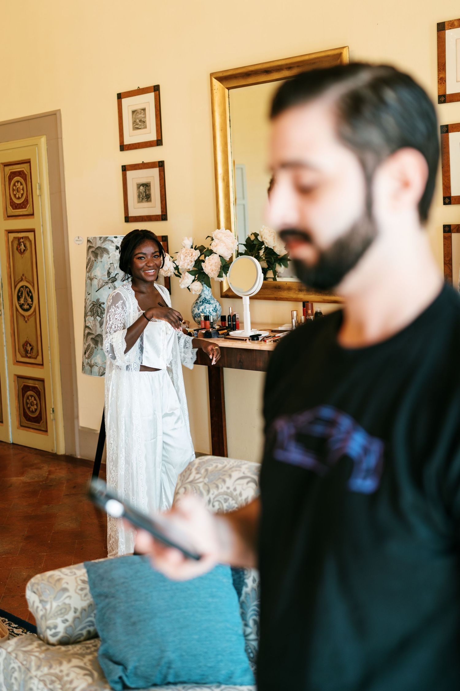 Smiling bride in a white lace robe stands by a vanity with flowers while a blurred bearded man checks his phone in the foreground.