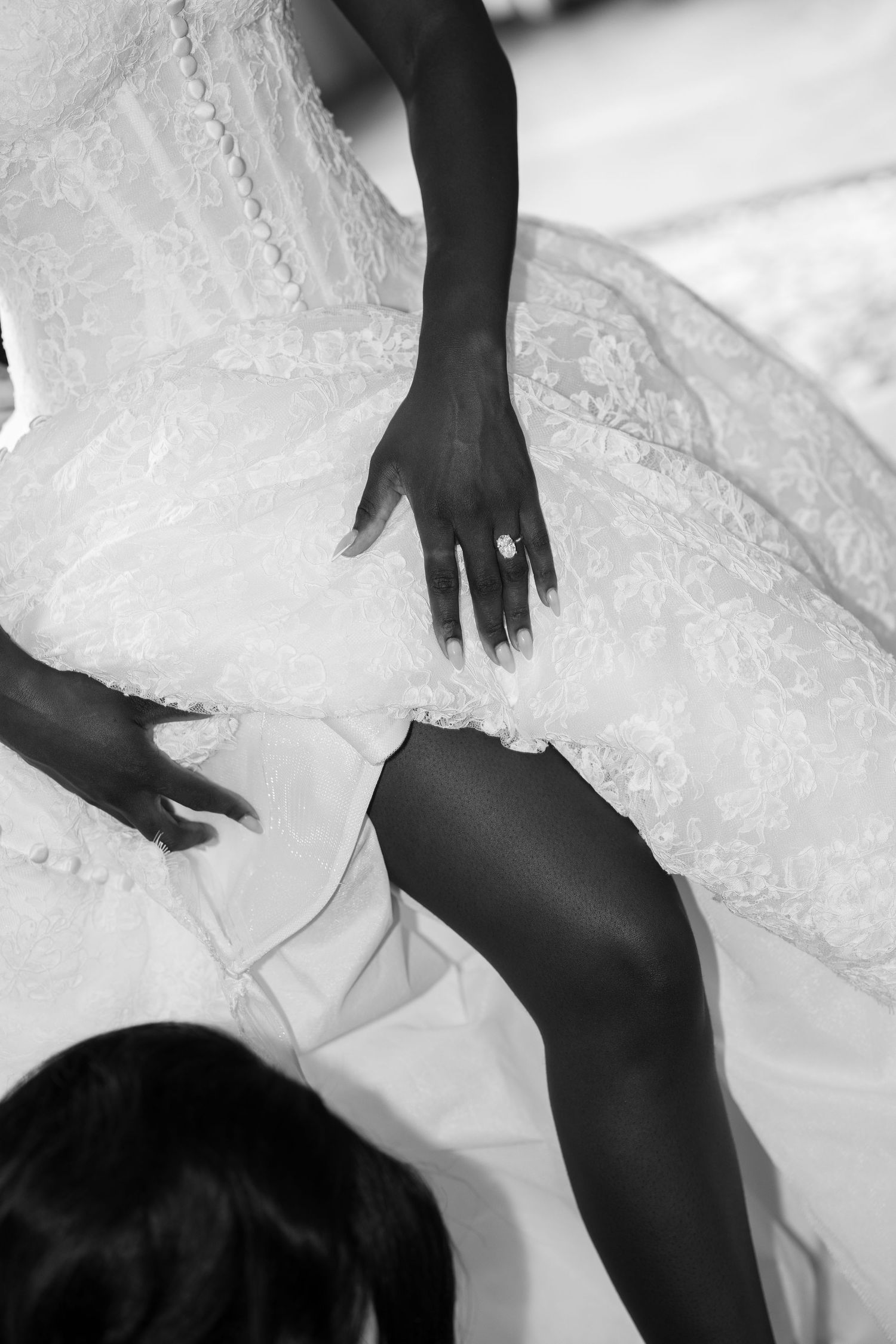 Black and white photo of a bride in a lace corset wedding gown with her ring hand on the skirt as a helper adjusts the dress.