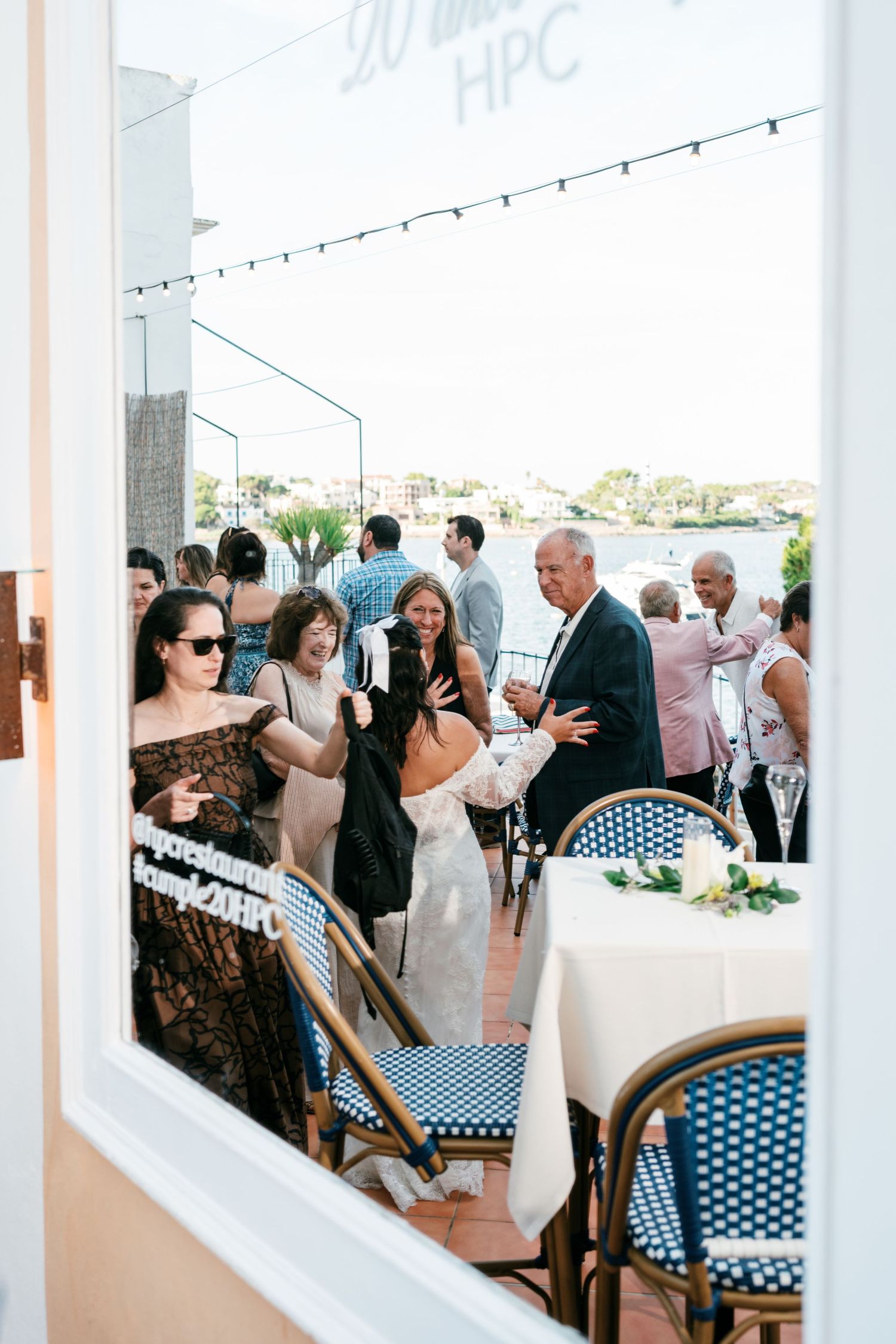 Wedding guests in elegant attire mingle and celebrate on a sunny seaside terrace with café chairs, a decorated table and waterfront views.