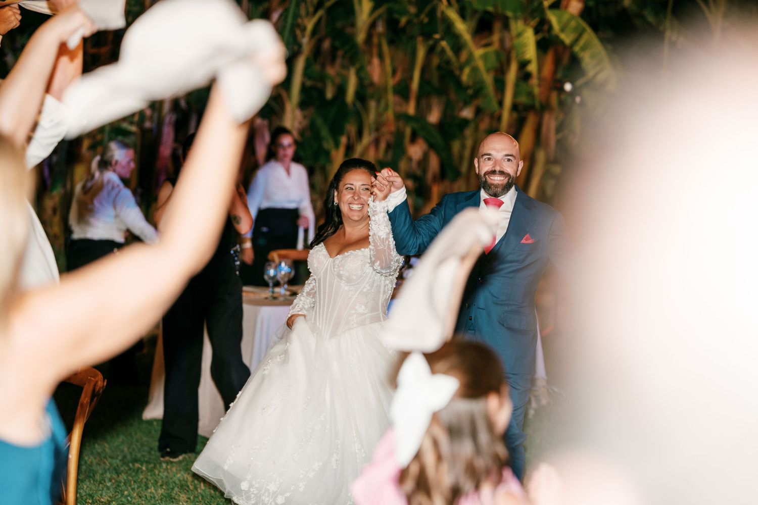 Bride in a white gown and groom in a navy suit raise their hands as wedding guests wave white napkins at an outdoor evening reception.