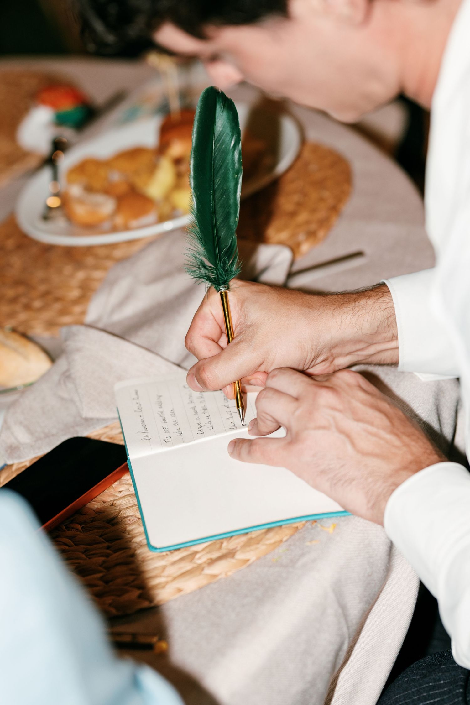 A guest in a white shirt writes a message in a guestbook with a green feather quill at a wedding reception table.