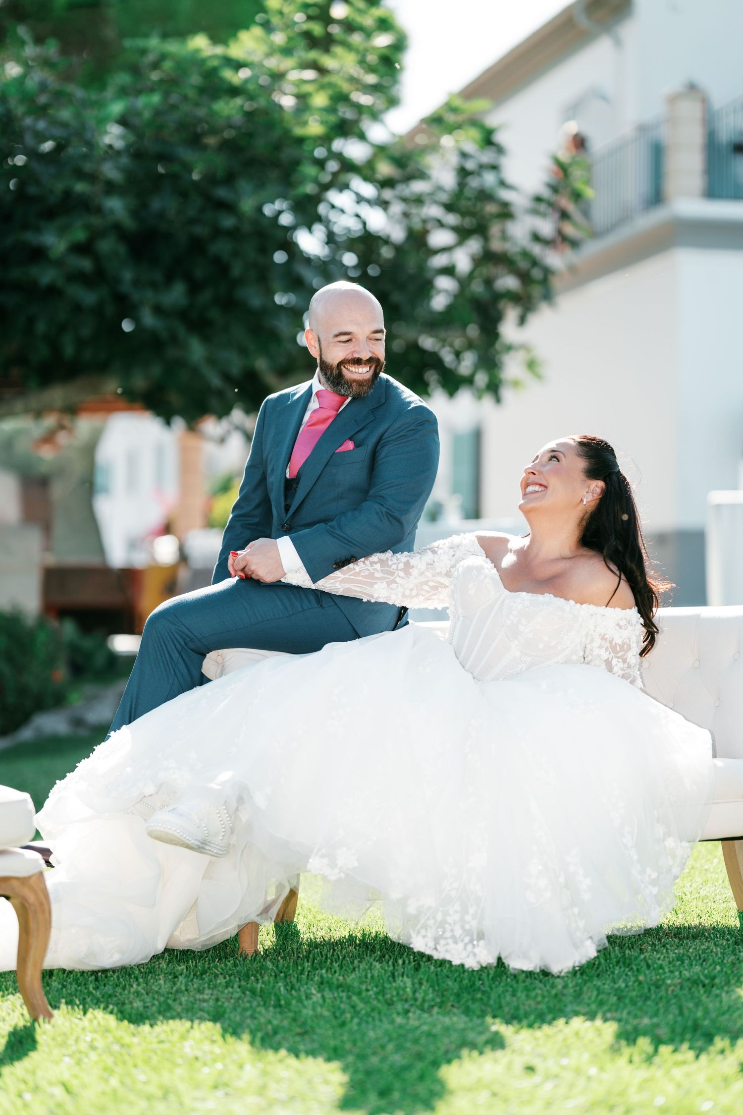 Smiling groom in a blue suit and pink tie sits beside a laughing bride in a lace tulle wedding gown on a white sofa outdoors on the lawn.