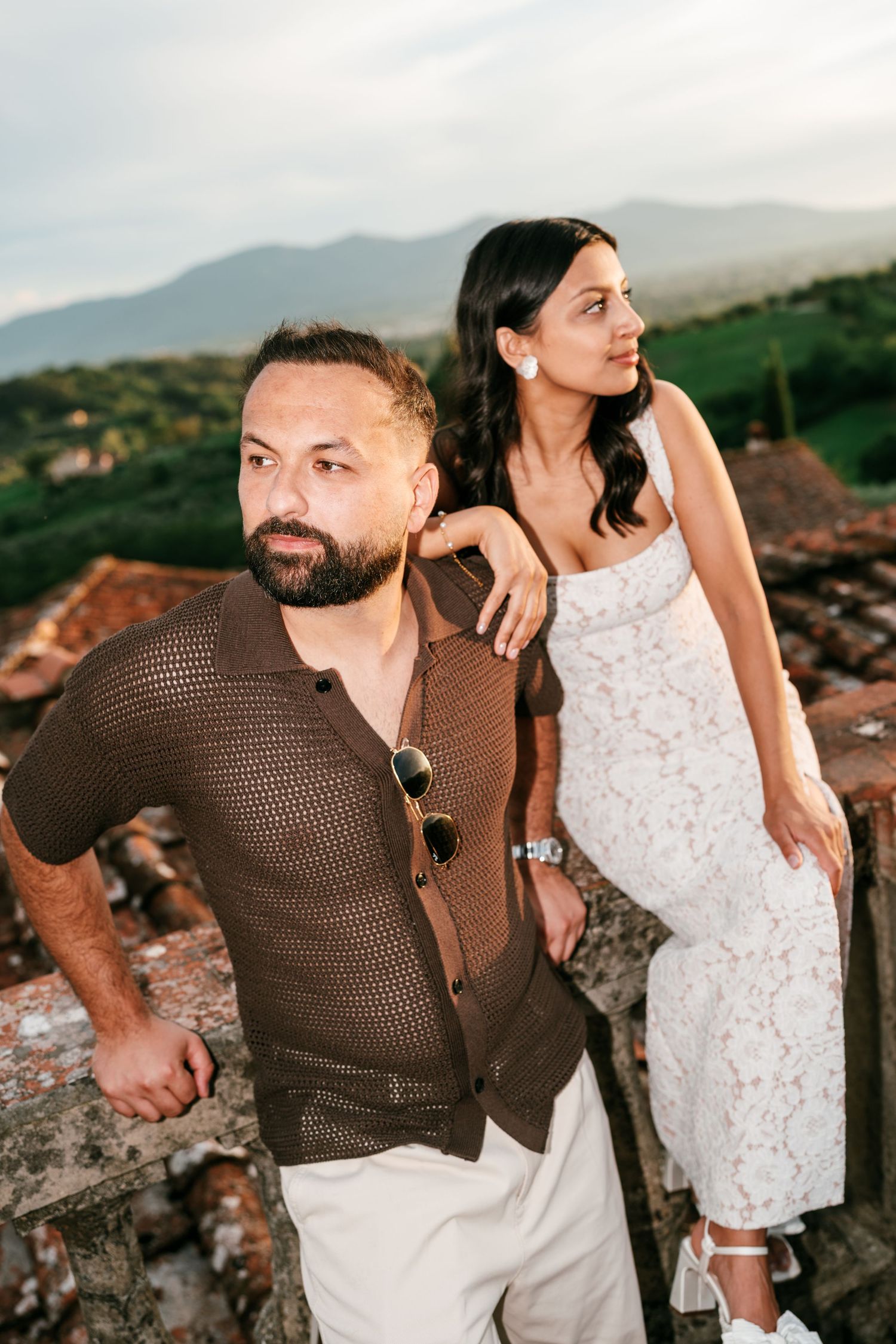 Couple posing on a rustic stone ledge overlooking green hills and mountains, he in a brown knit shirt, she in a white lace dress.