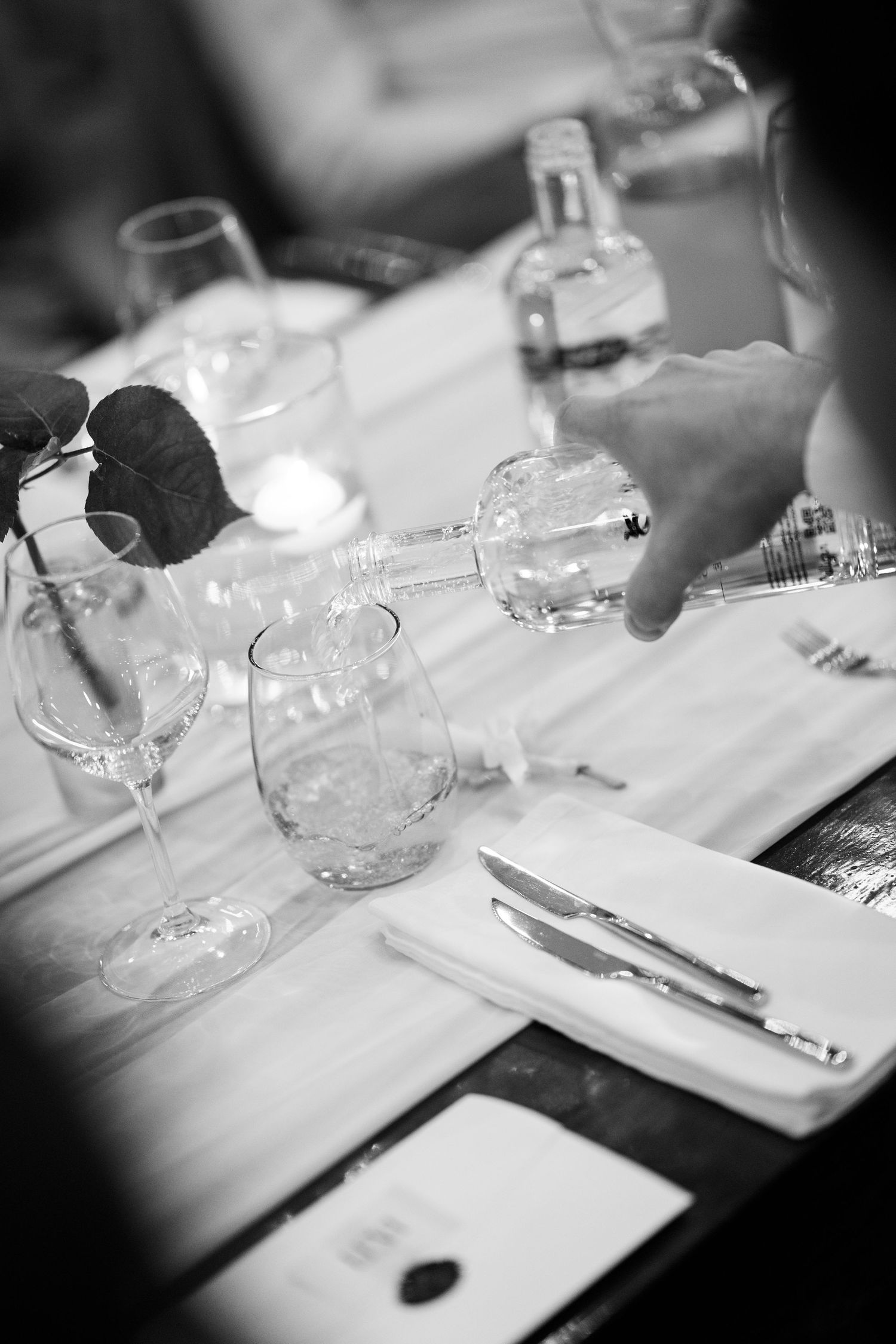 Black and white photo of elegant table setting with wine glasses and a hand pouring water at a formal dinner event.