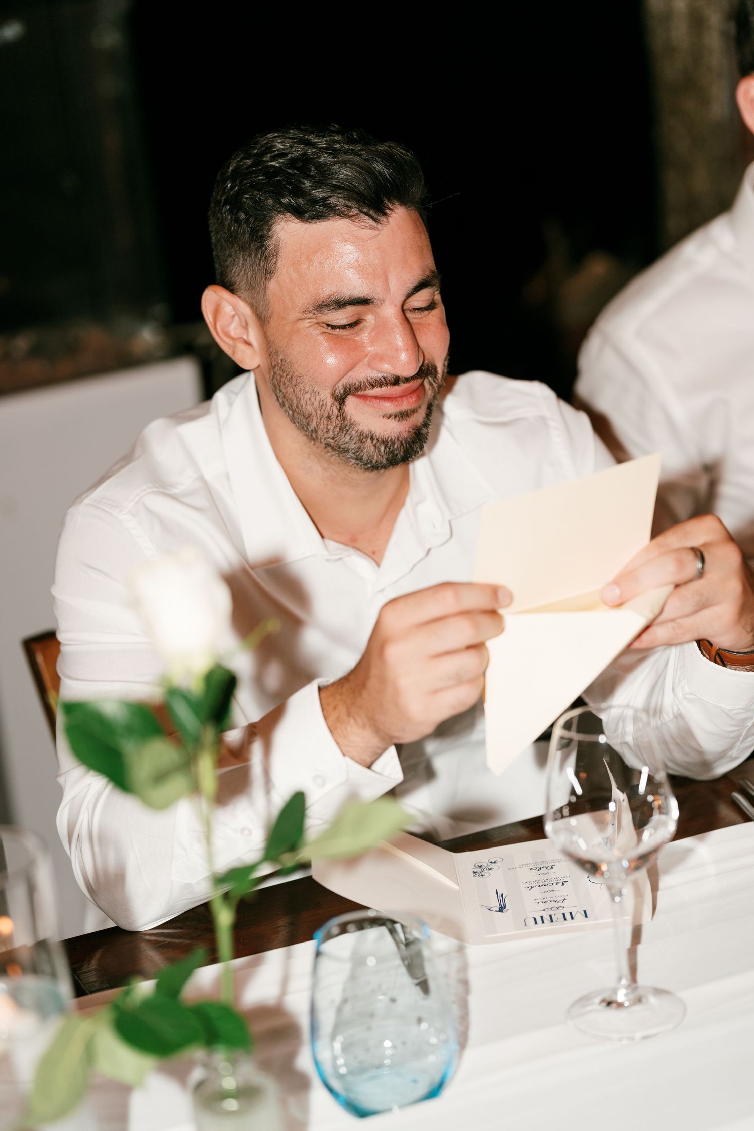 A smiling bearded man in a white shirt reads a card at an elegant outdoor wedding reception dinner table.