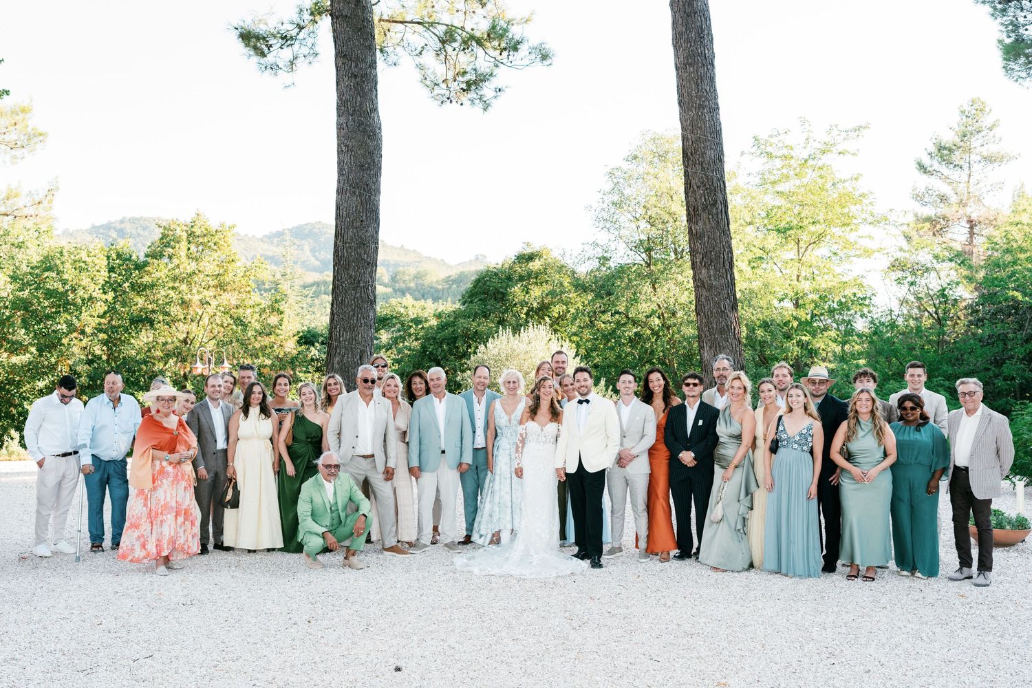 Large wedding group photo outdoors with guests in elegant attire, surrounded by tall trees and lush greenery.