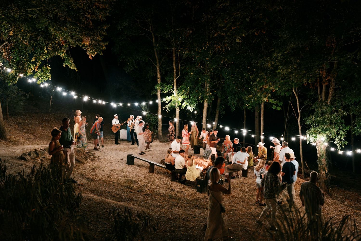 Outdoor evening gathering with string lights among trees, guests mingling around tables on sandy ground at night.