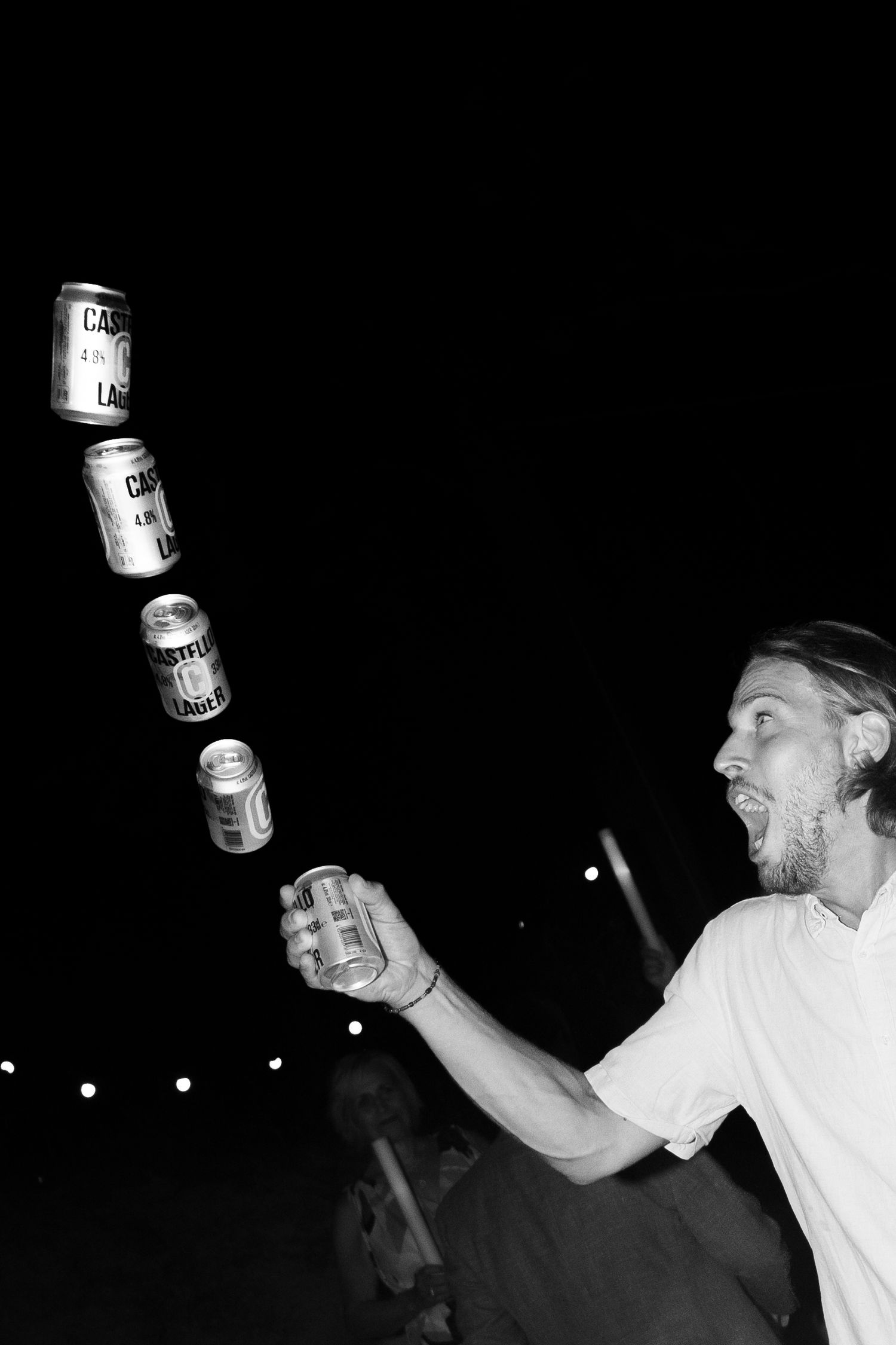 Black and white photo of a man tossing multiple Castle Lager beer cans in the air at a night event.
