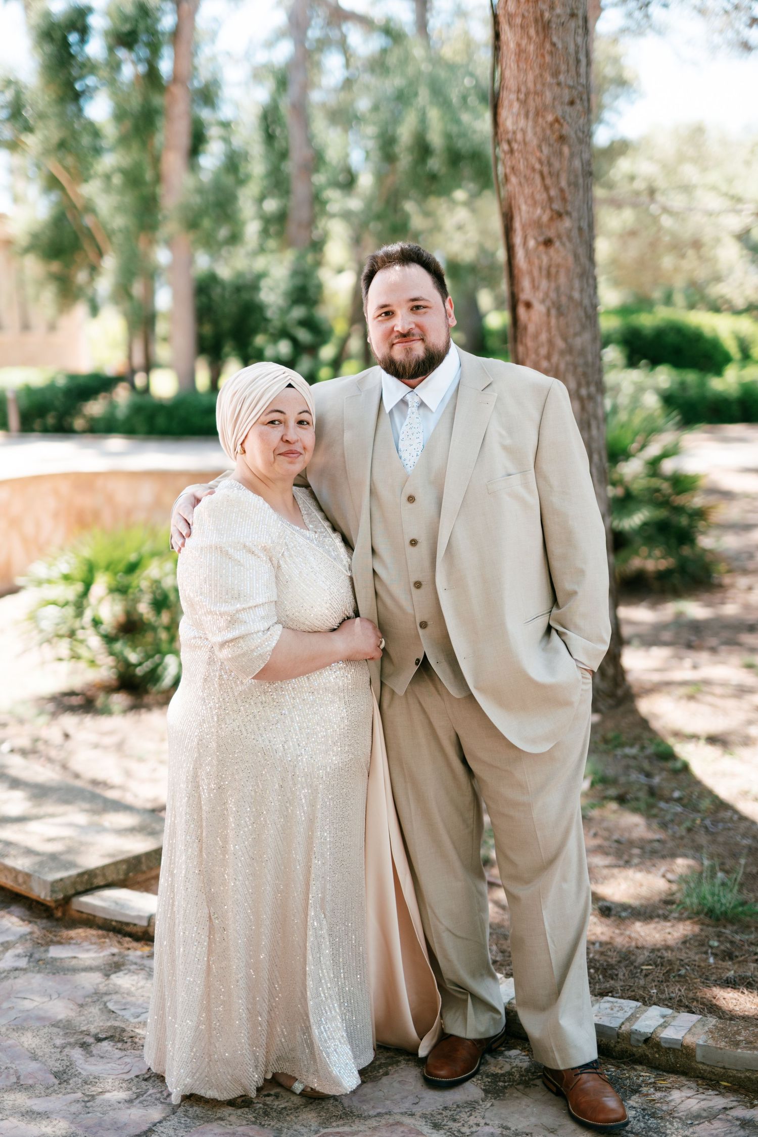 Couple in formal attire posing outdoors, woman in sparkly dress with head covering, man in tan suit, among trees in Mallorca.