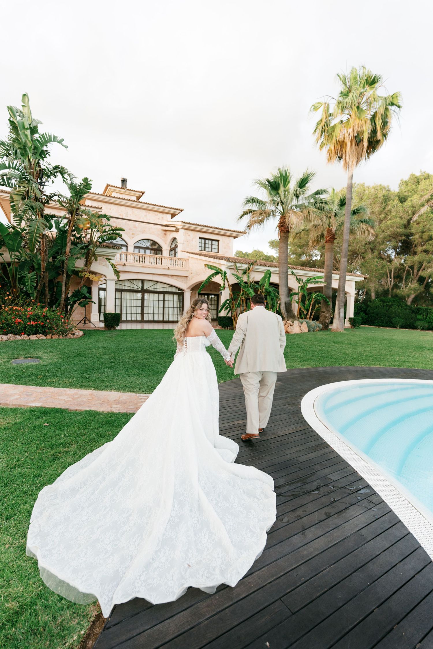Bride and groom walking hand in hand along a wooden deck beside a pool at a luxury villa wedding venue.