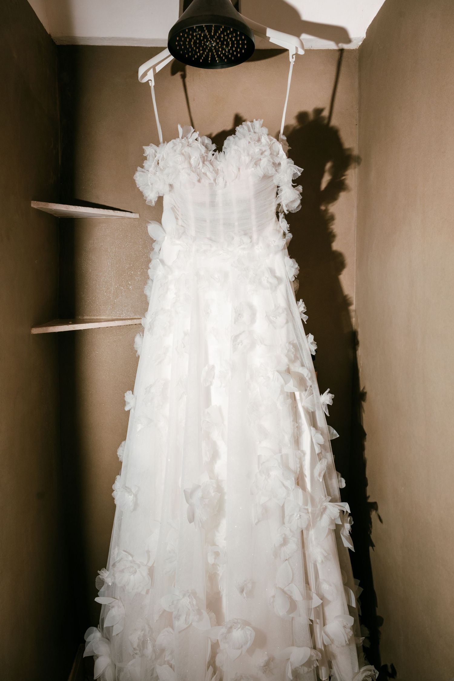 A stunning white wedding dress with 3D floral details hanging in a warmly lit closet on a hanger. Mallorca wedding photographer, filmmaker