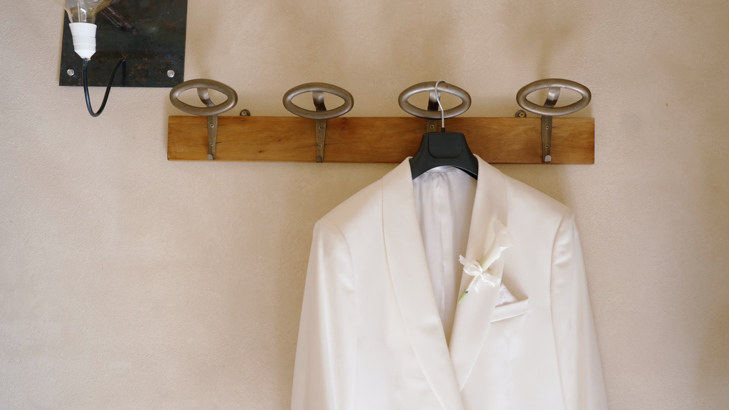 White dinner jacket with a boutonniere hanging on a black hanger from a rustic wooden wall rack with round metal hooks.