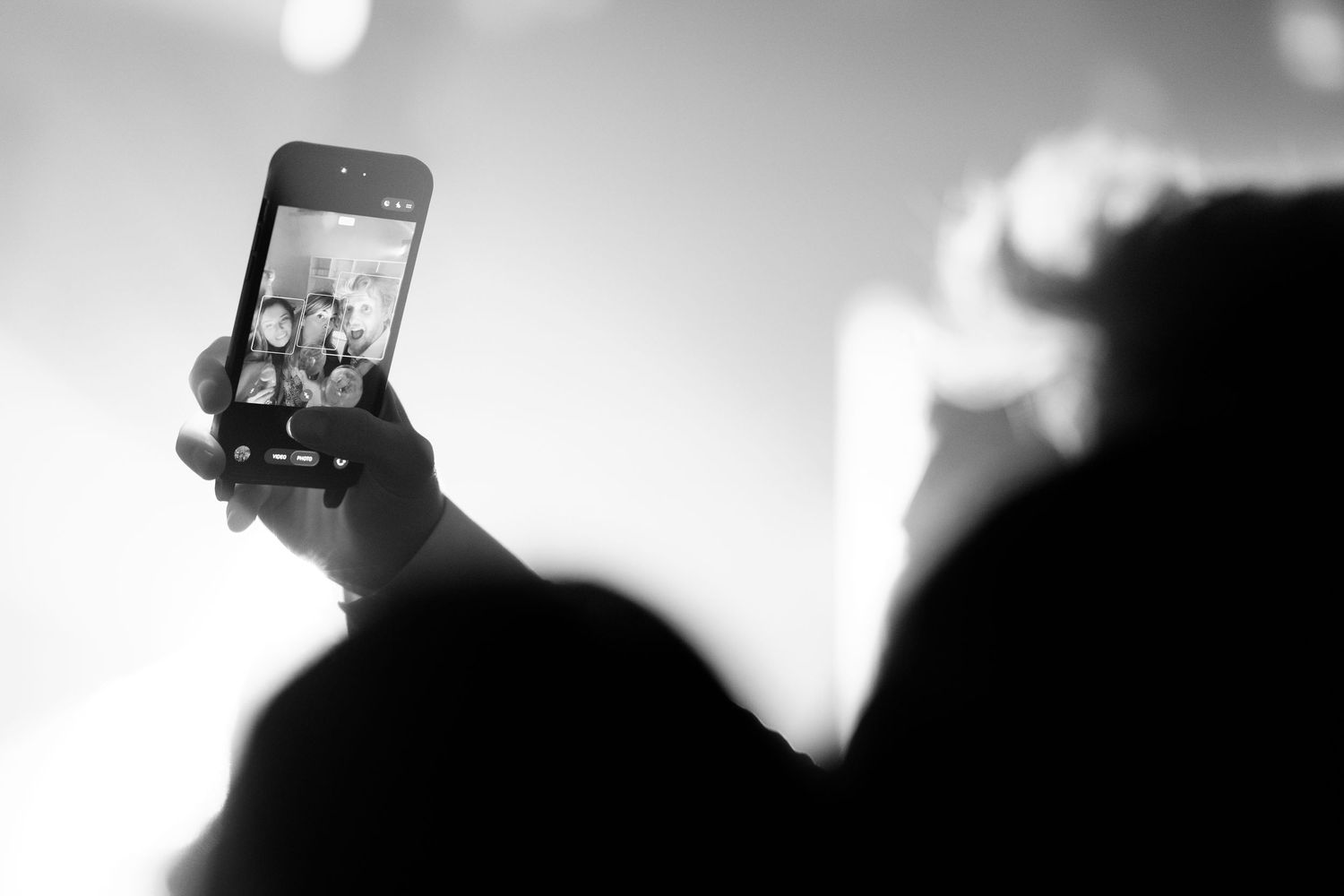 Black and white photo of a hand holding a smartphone to capture a group photo on its screen at a crowded, softly lit concert.
