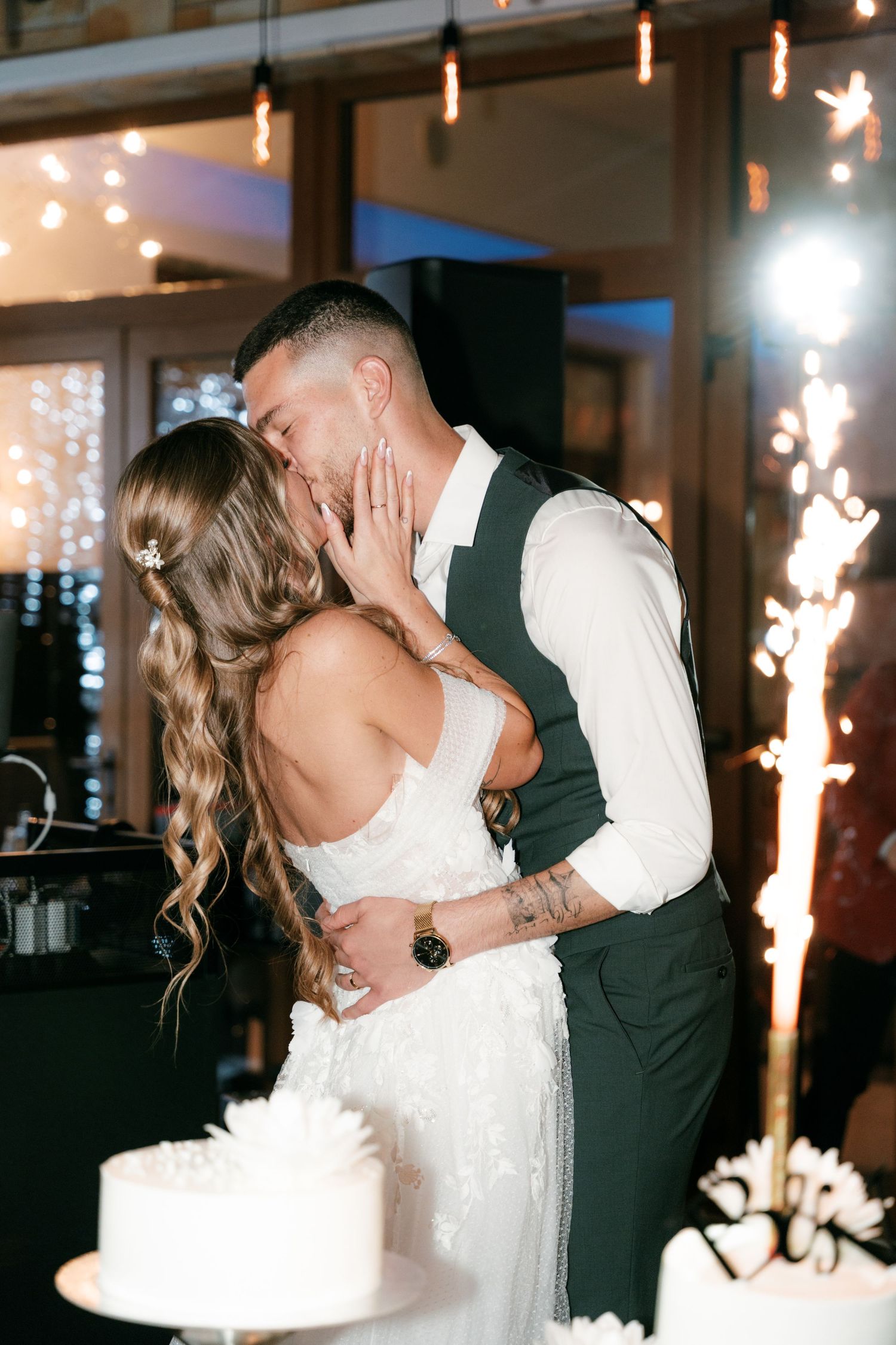 Bride and groom share a romantic kiss beside their white wedding cake with sparklers glowing in the background. Mallorca wedding photographe