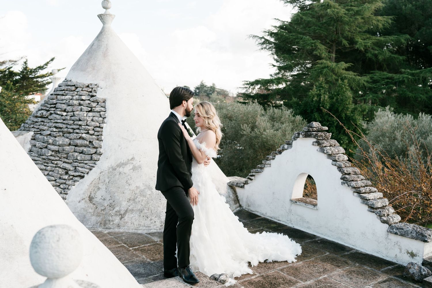 Bride and groom sharing a romantic kiss atop white trulli rooftops surrounded by cypress trees. Puglia wedding photographer, film