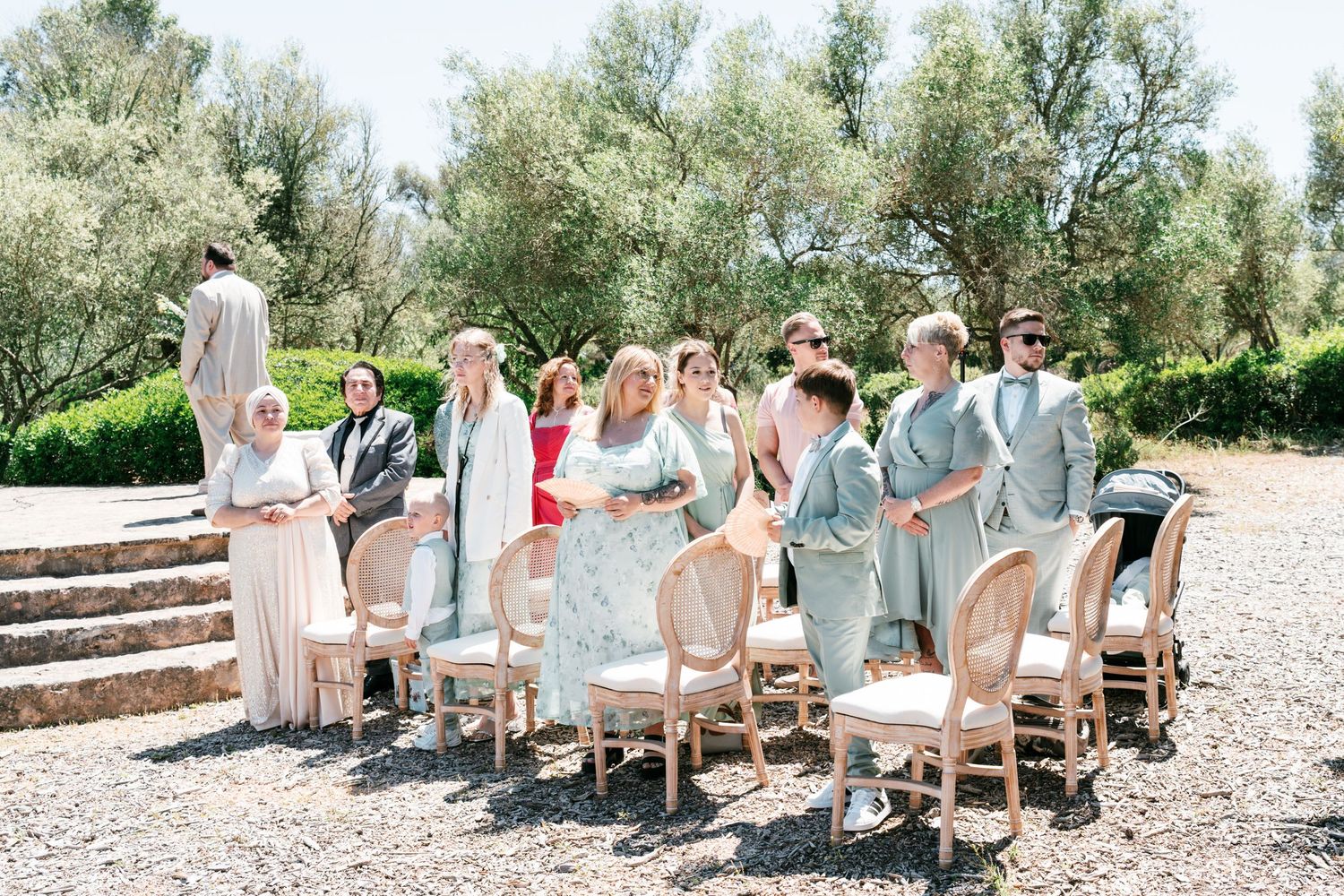 Guests in pastel attire stand and mingle at an outdoor wedding ceremony surrounded by olive trees on a sunny day in Mallorca.