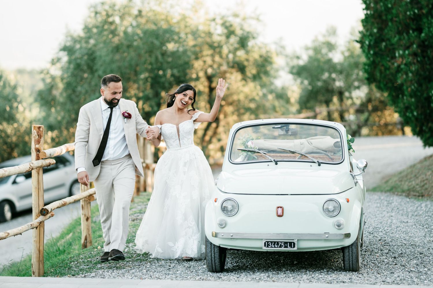 Bride in a white gown and groom in a light suit walk hand in hand beside a vintage white Fiat 500 on a scenic gravel path.