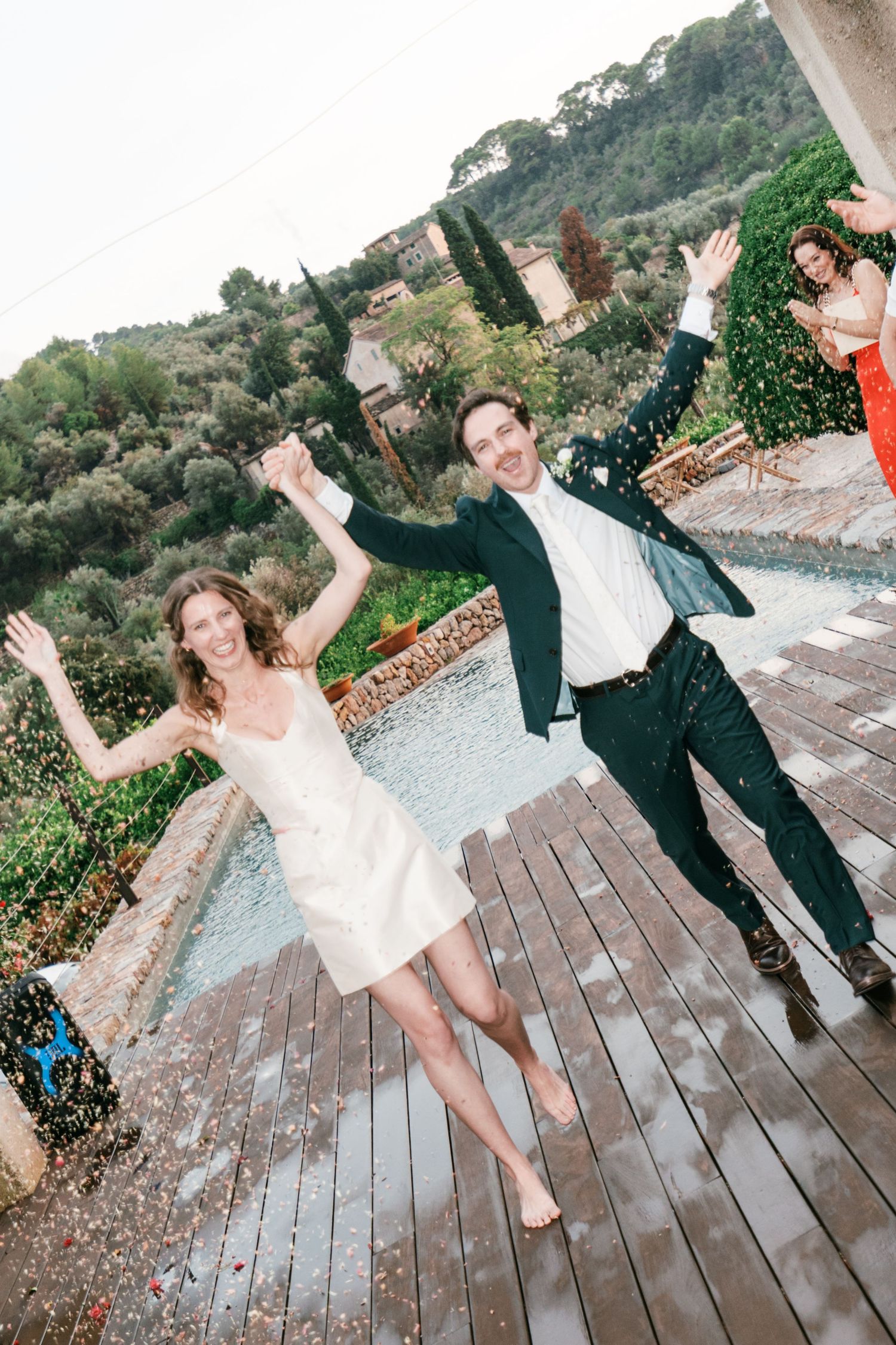 Joyful newlyweds dance barefoot on a wet wooden deck beside a pool, celebrating with raised arms amid confetti and a scenic hillside.
