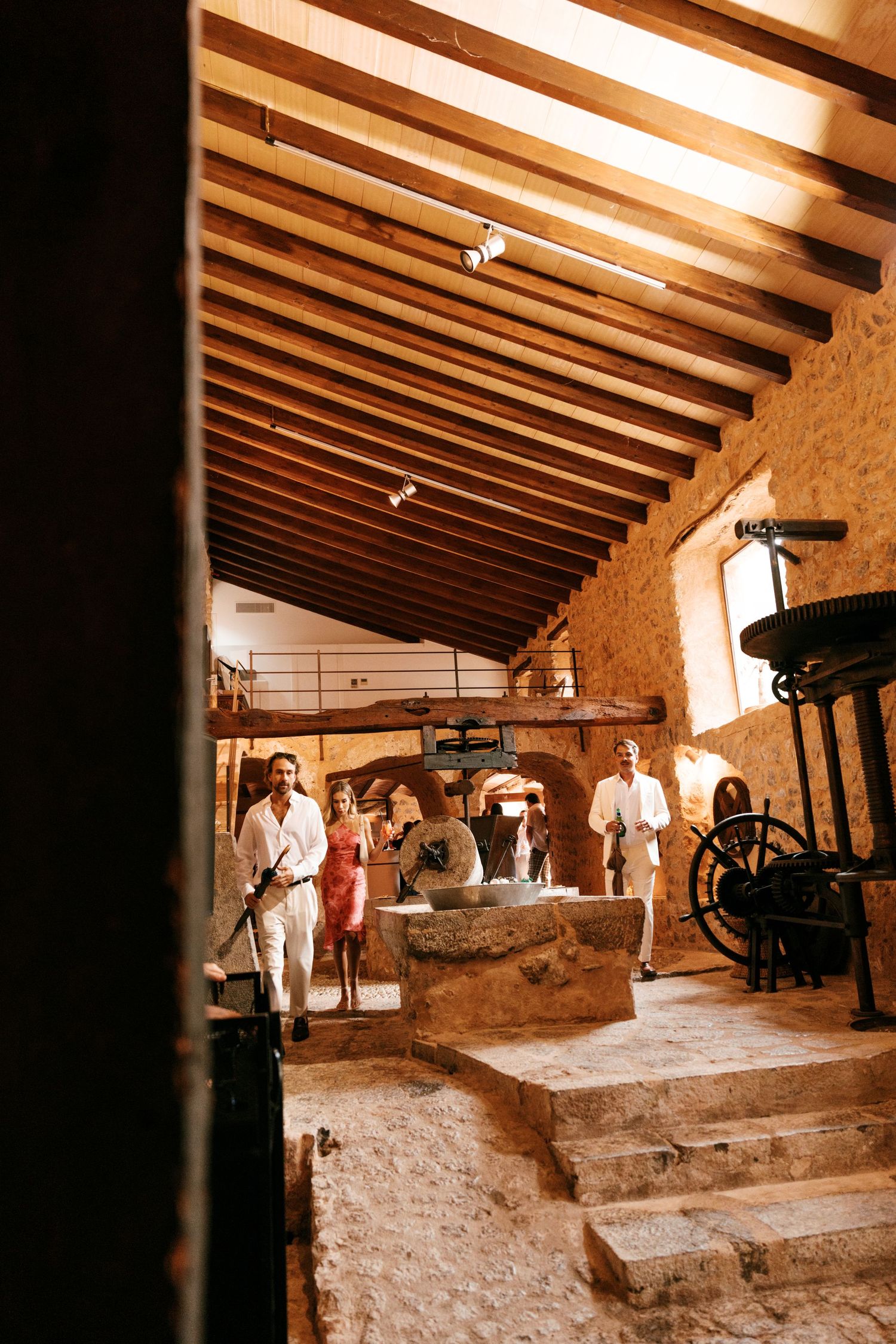 Wedding guests walk through a rustic stone mill with exposed wooden beams, vintage machinery, and a sunlit rough-hewn interior.