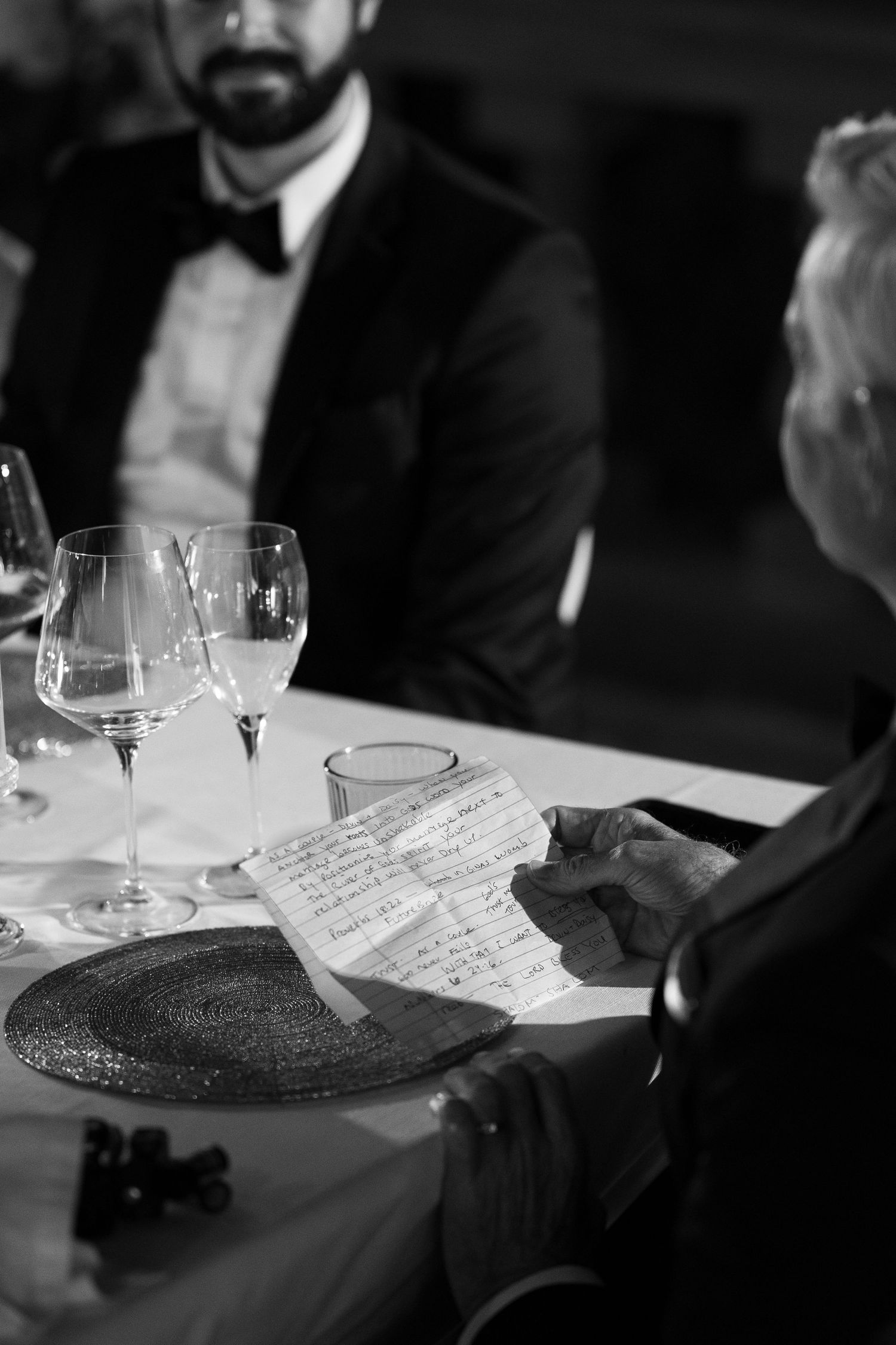 Black and white photo of a guest reading a handwritten note at a dinner table with wine glasses while a man in a bow tie looks on.