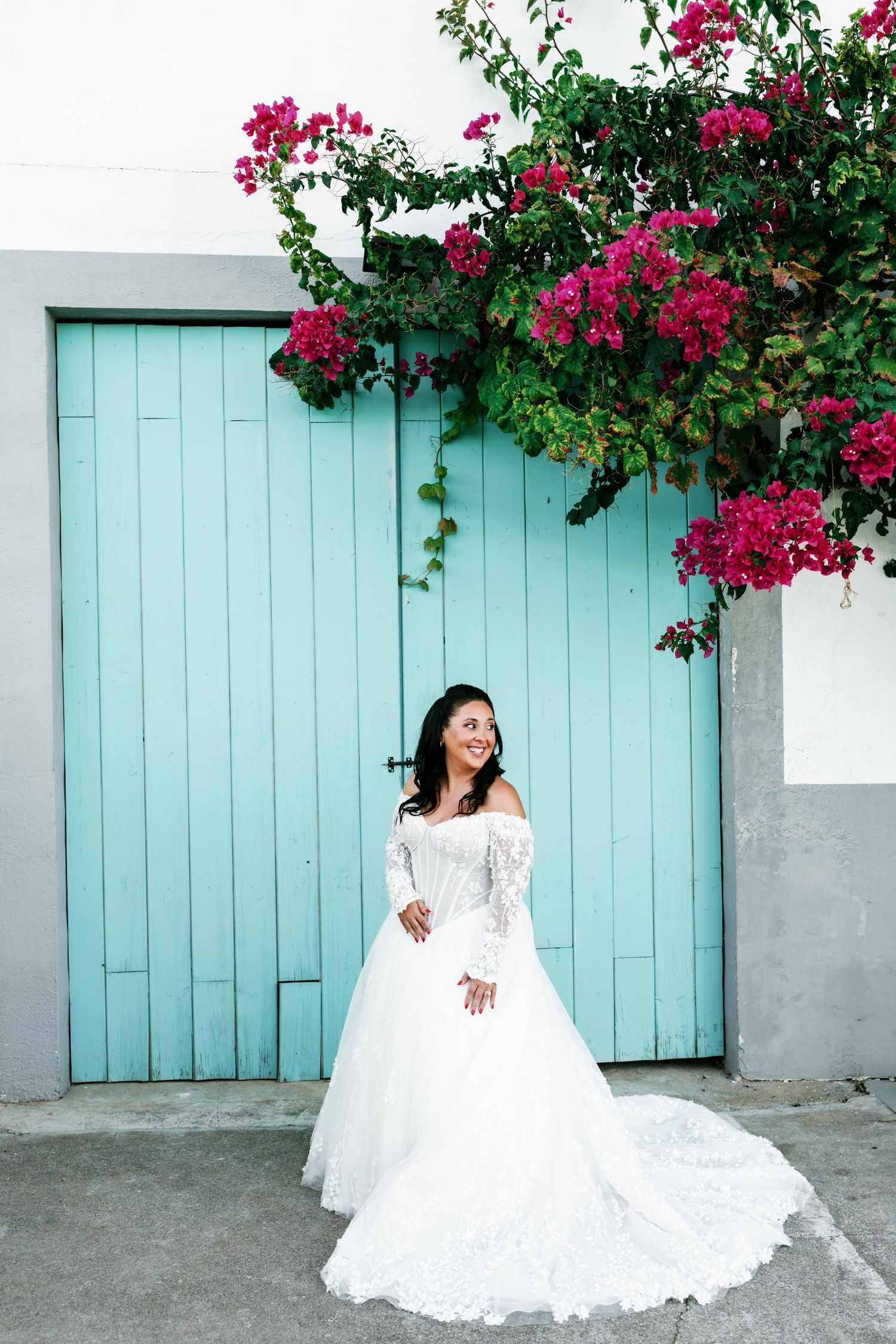 Smiling bride in a white lace off-the-shoulder wedding gown poses before a turquoise wooden door beneath pink bougainvillea.