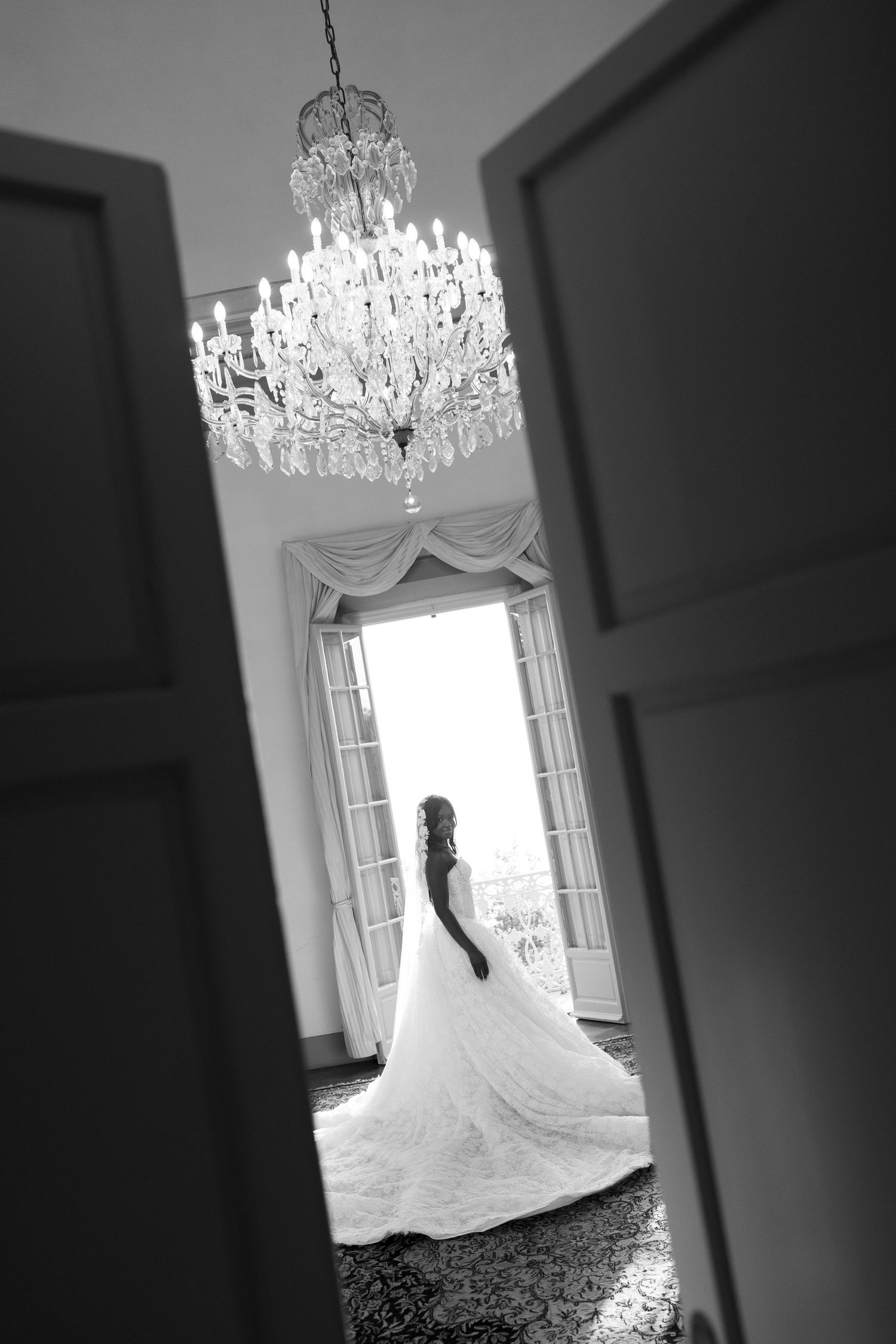 Black and white photo of a bride in a flowing gown standing before open doors beneath a crystal chandelier.