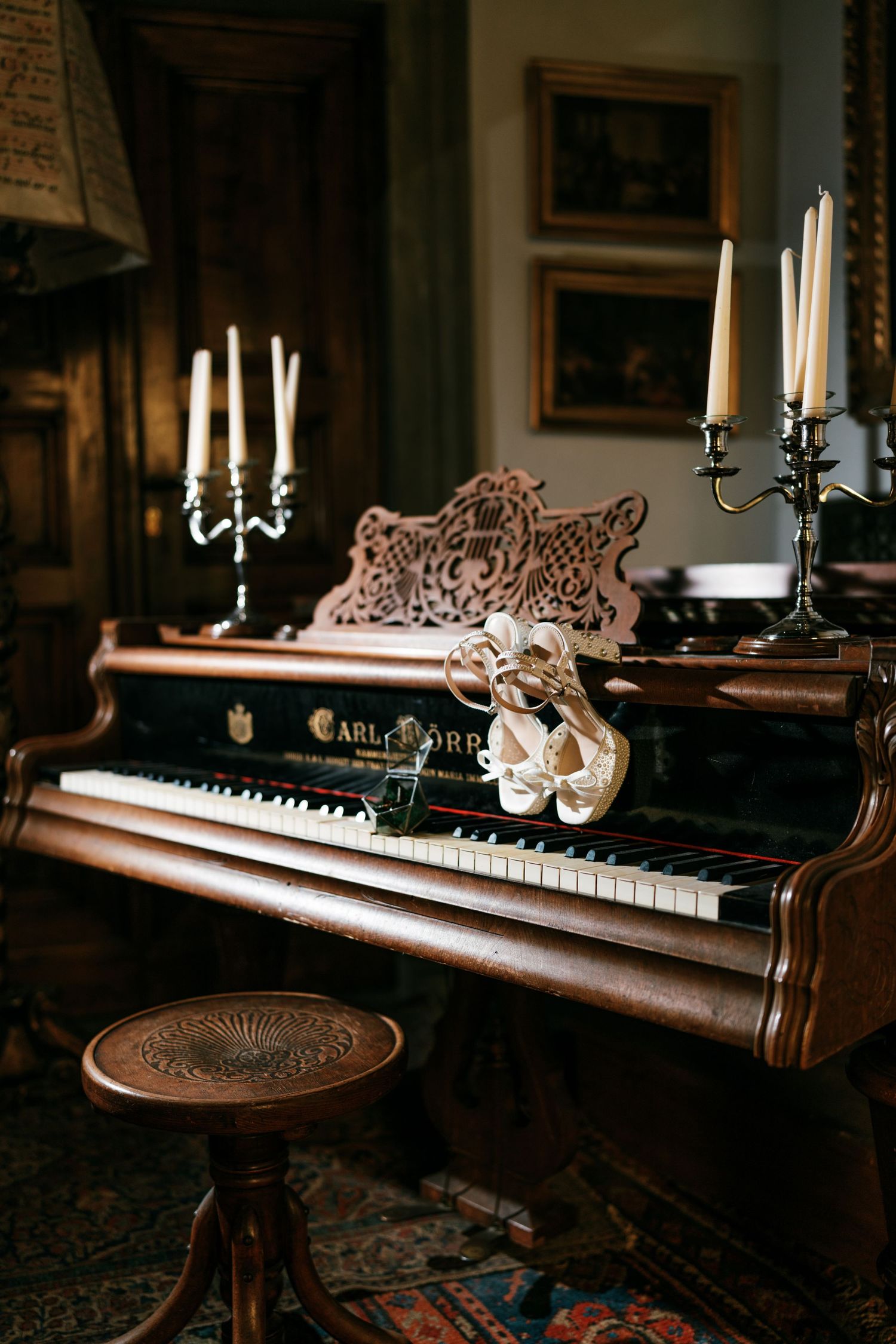 Antique upright piano with candelabras and a pair of ivory bridal heels resting on the keys beside a carved wooden stool in a vintage room.