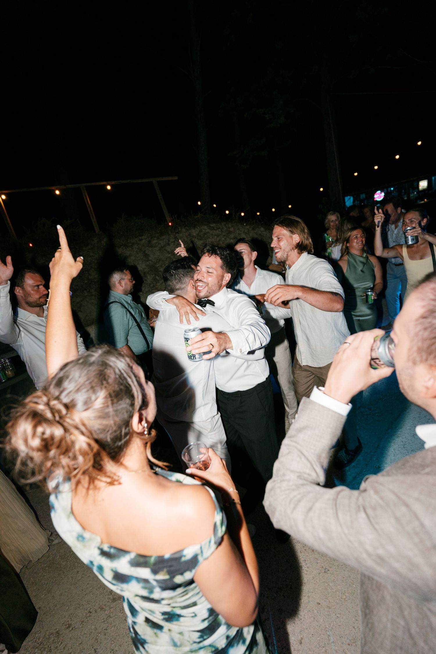 Guests dancing and embracing joyfully at an outdoor evening wedding reception under string lights.
