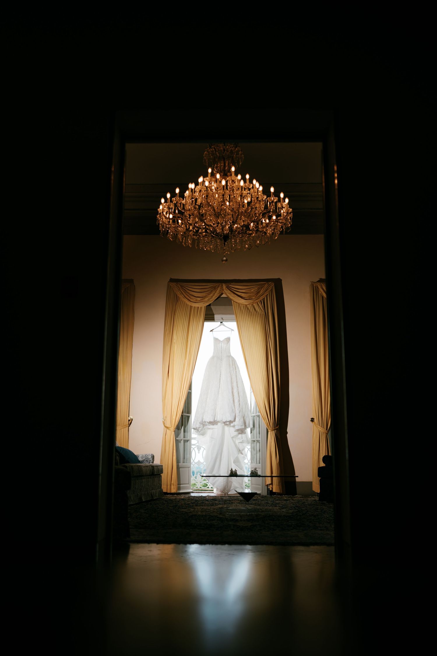 White wedding dress hanging in a tall window with gold curtains beneath an ornate crystal chandelier in an elegant, dimly lit room.