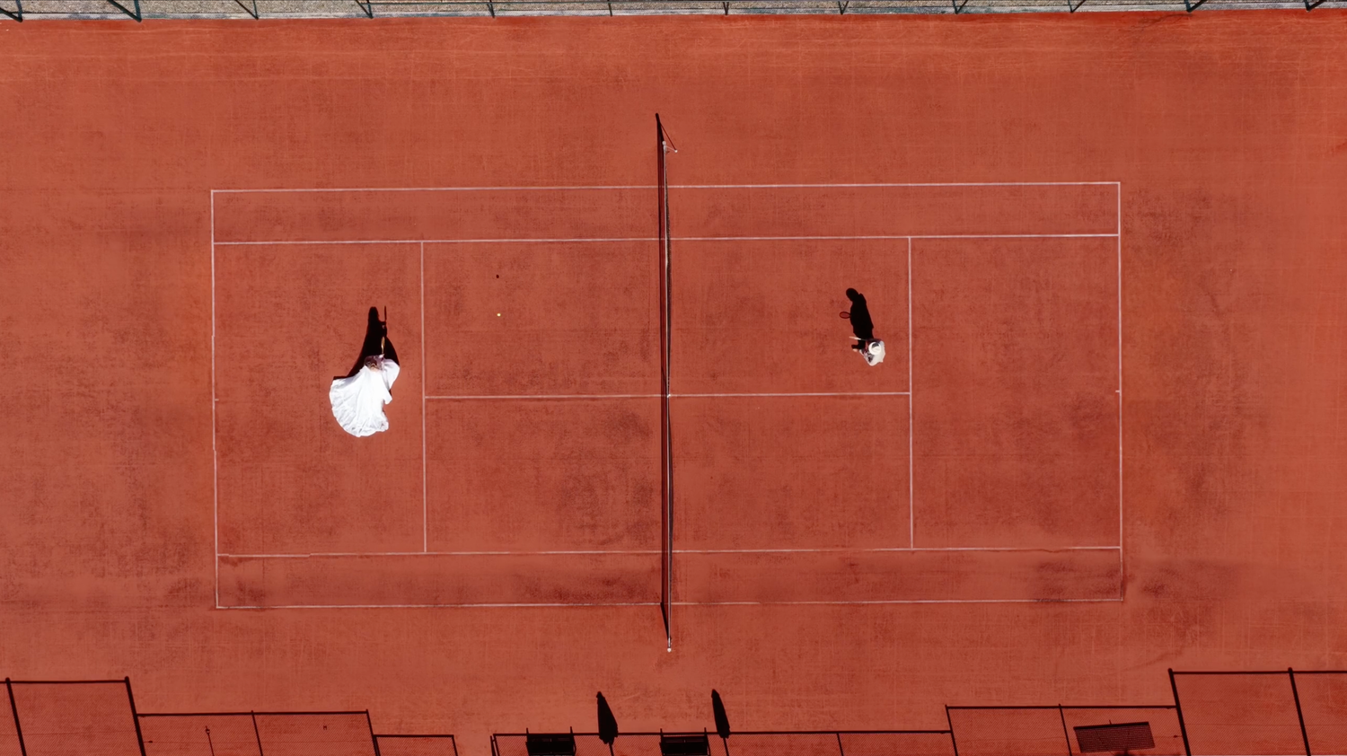 Aerial view of a bride in a white wedding dress and a groom in dark attire standing on opposite sides of a red clay tennis court.