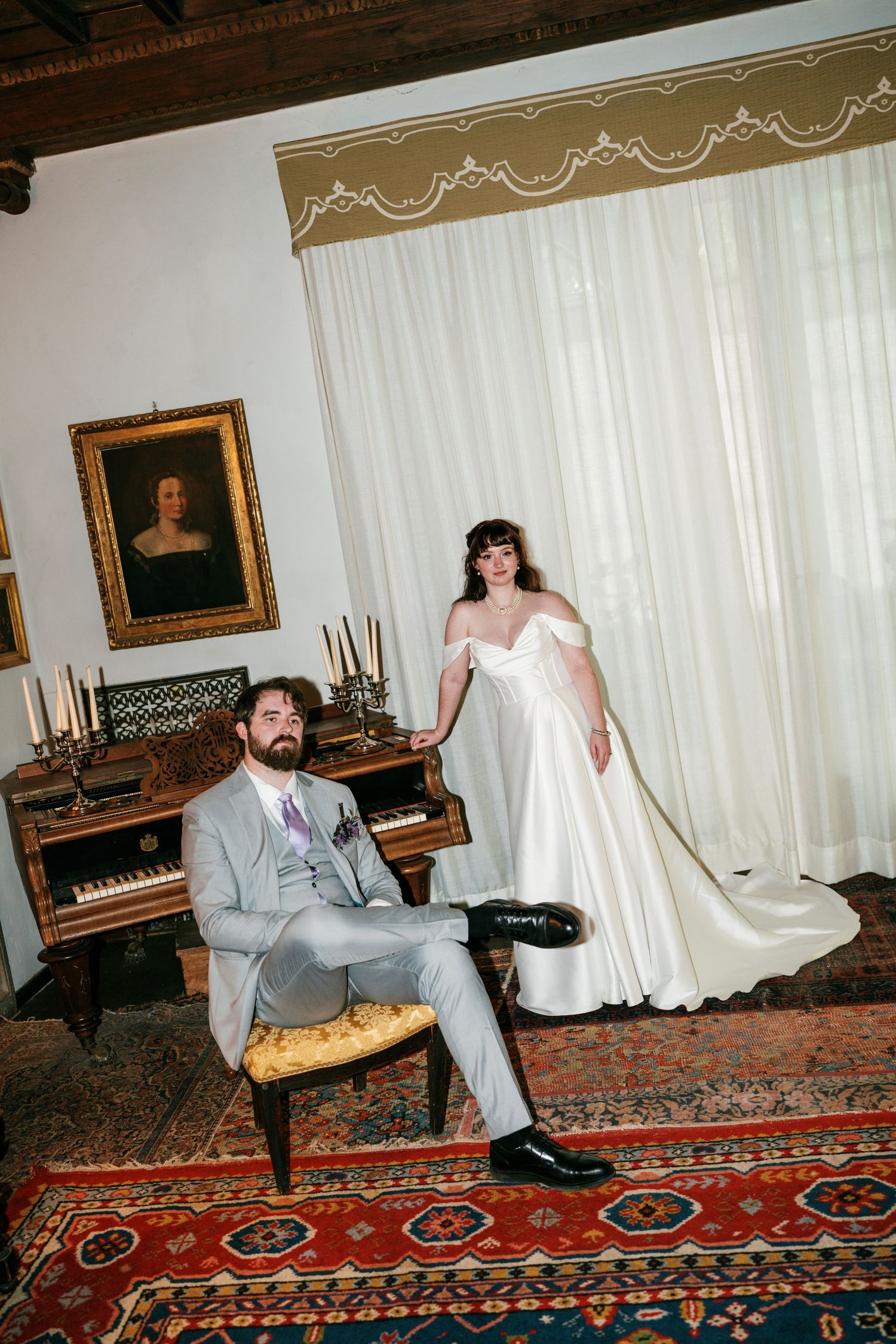 Bride in off-shoulder gown stands beside groom in gray suit seated at antique piano in elegant room.