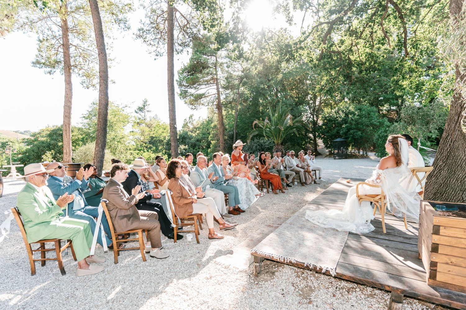 Guests seated outdoors at a sunny wedding ceremony surrounded by tall trees and dappled light.
