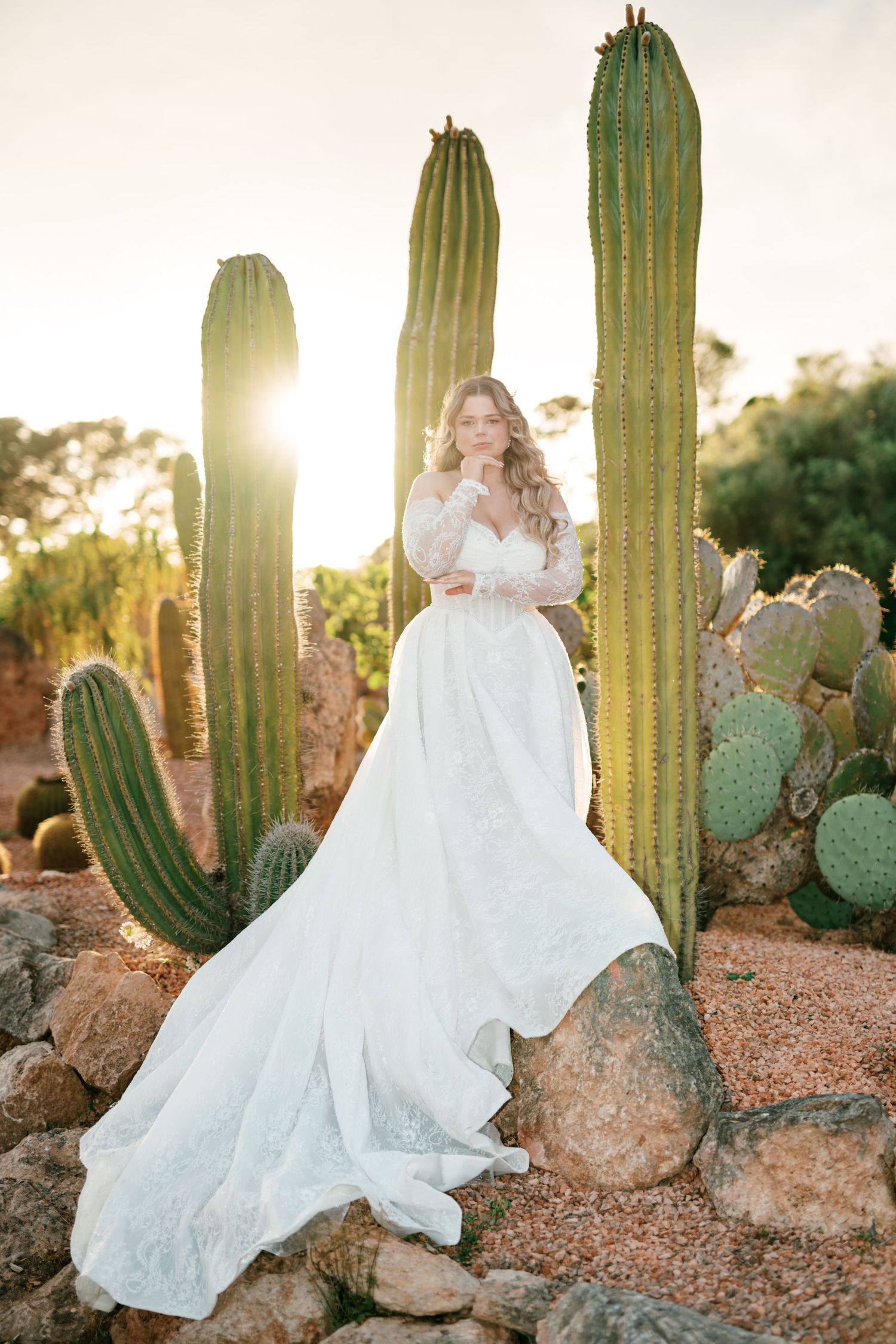 Bride in flowing white gown posing among tall cacti at golden sunset in desert garden in Mallorca.