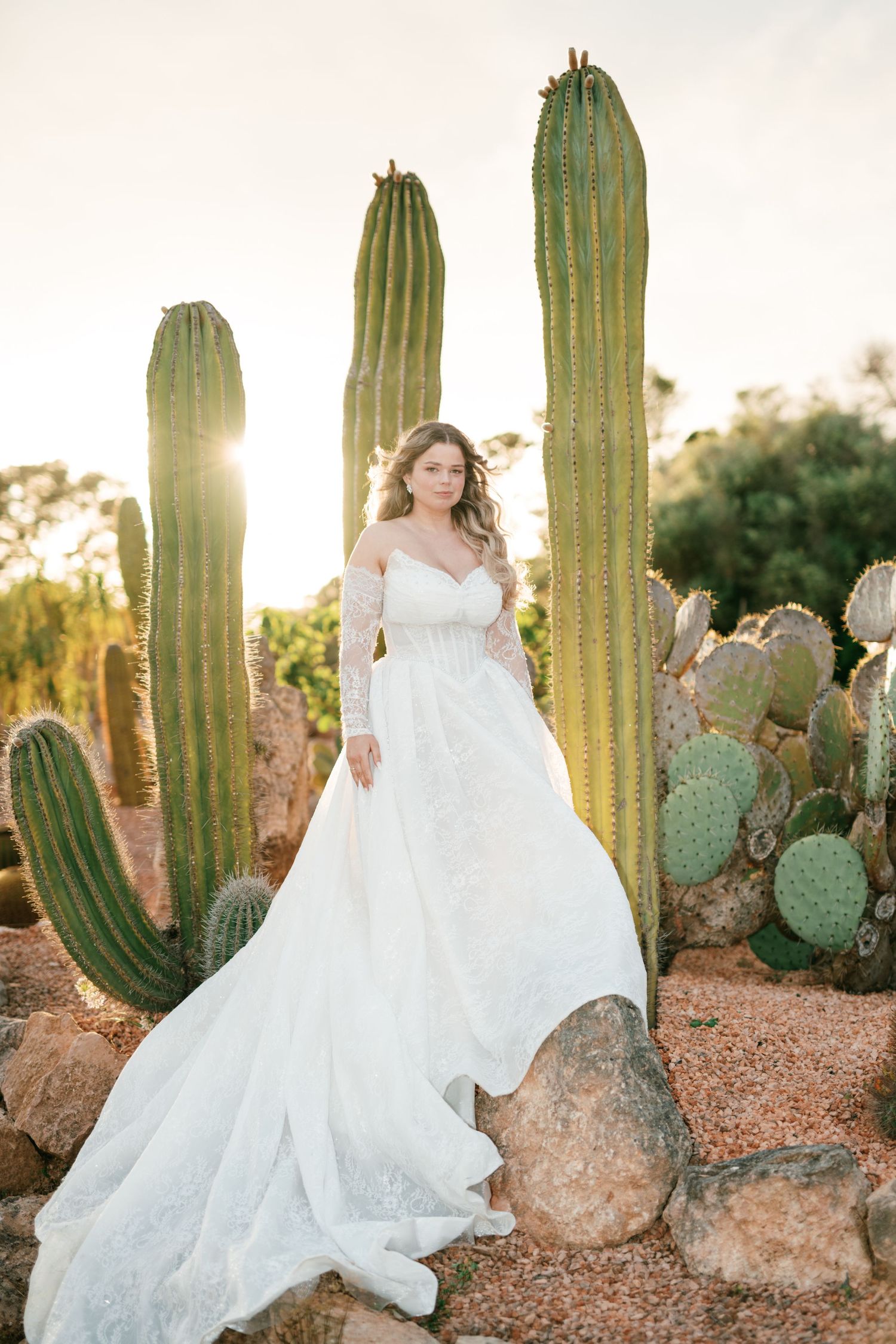 Bride in white lace off-shoulder gown with long train poses among tall saguaro cacti at golden hour.