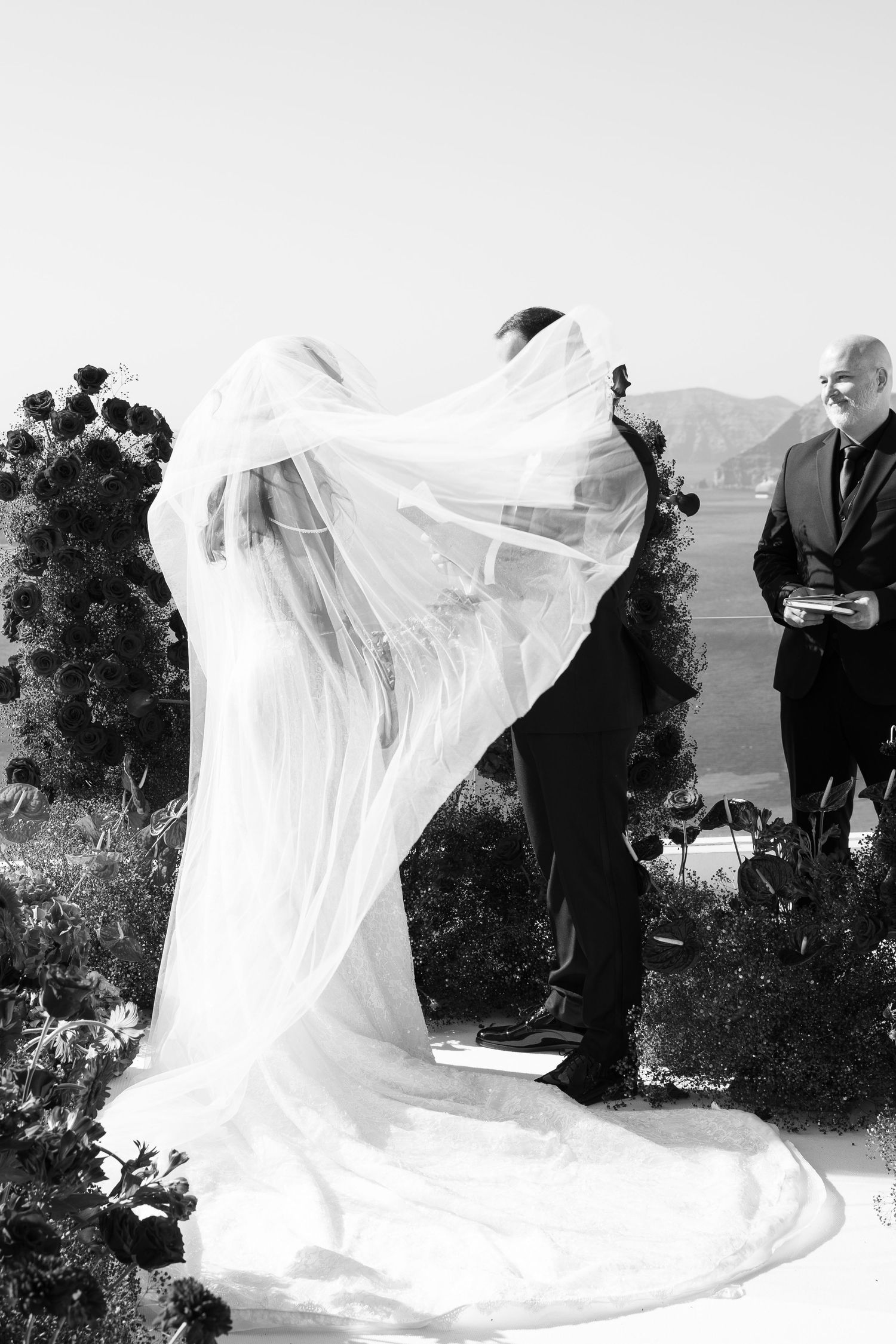 Black and white photo of bride and groom kissing as her veil billows in the wind during outdoor wedding ceremony.