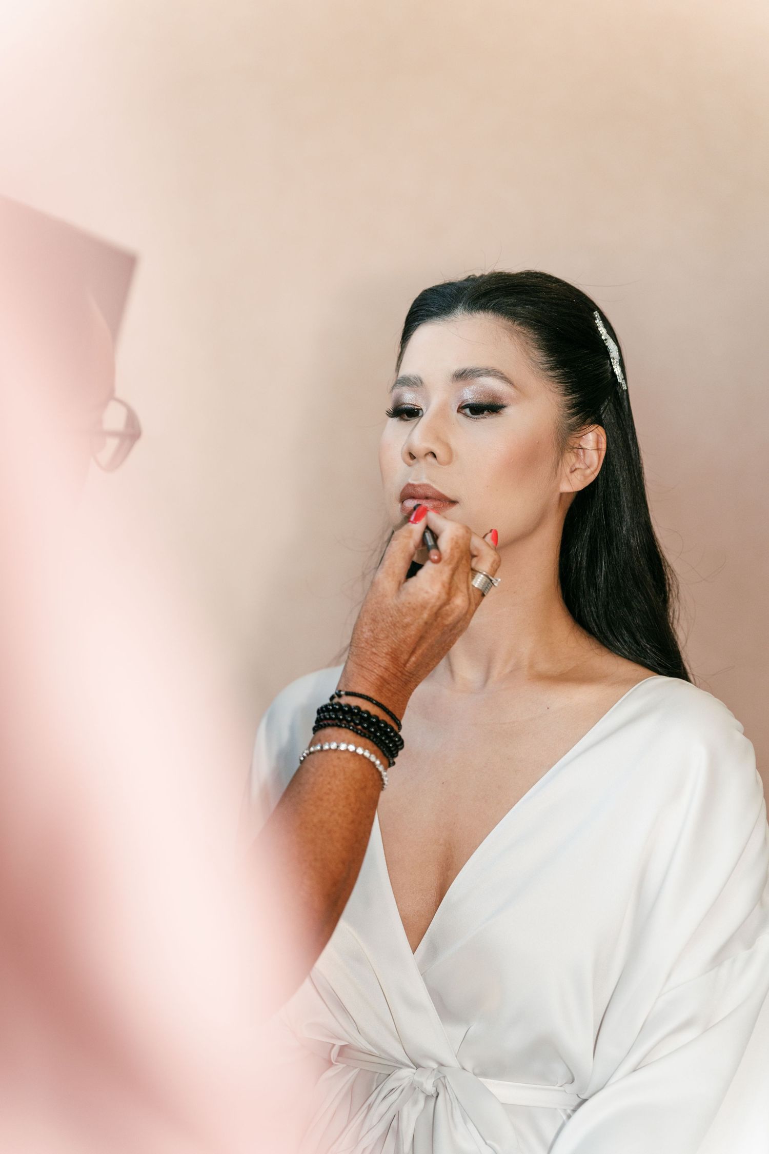 Makeup artist applying red lipstick to a bride in a white robe with dark hair and a hair accessory. Italy wedding photographer and filmmaker